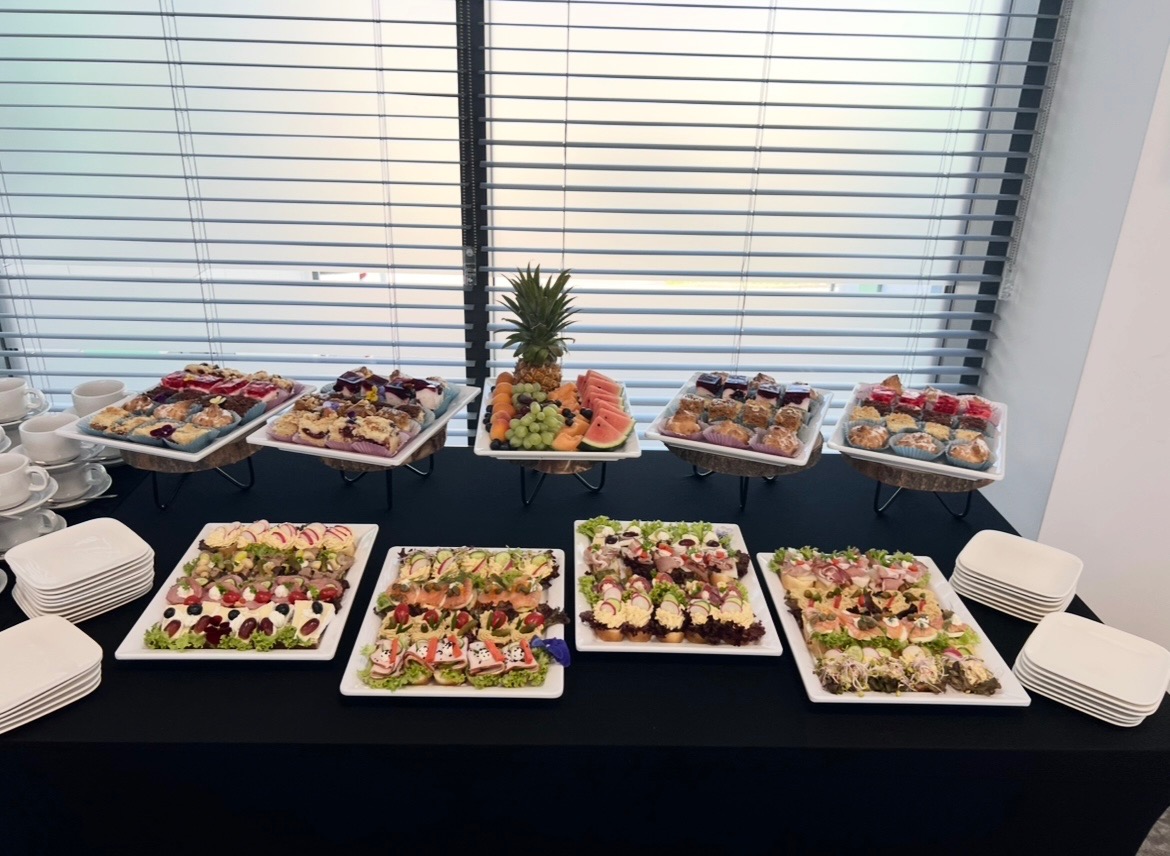 Stół cateringowy z przekąskami: kanapeczki, ciasta, owoce (ananas na środku), białe talerze, filiżanki. Rolety w tle, jasne wnętrze.