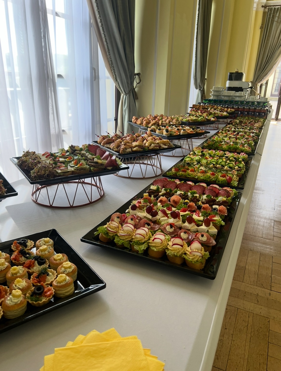Długi stół z różnorodnymi, apetycznie ułożonymi przekąskami na czarnych tacach, idealny na catering firmowy lub event. Widoczne kanapki, tartaletki i mini-croissanty.