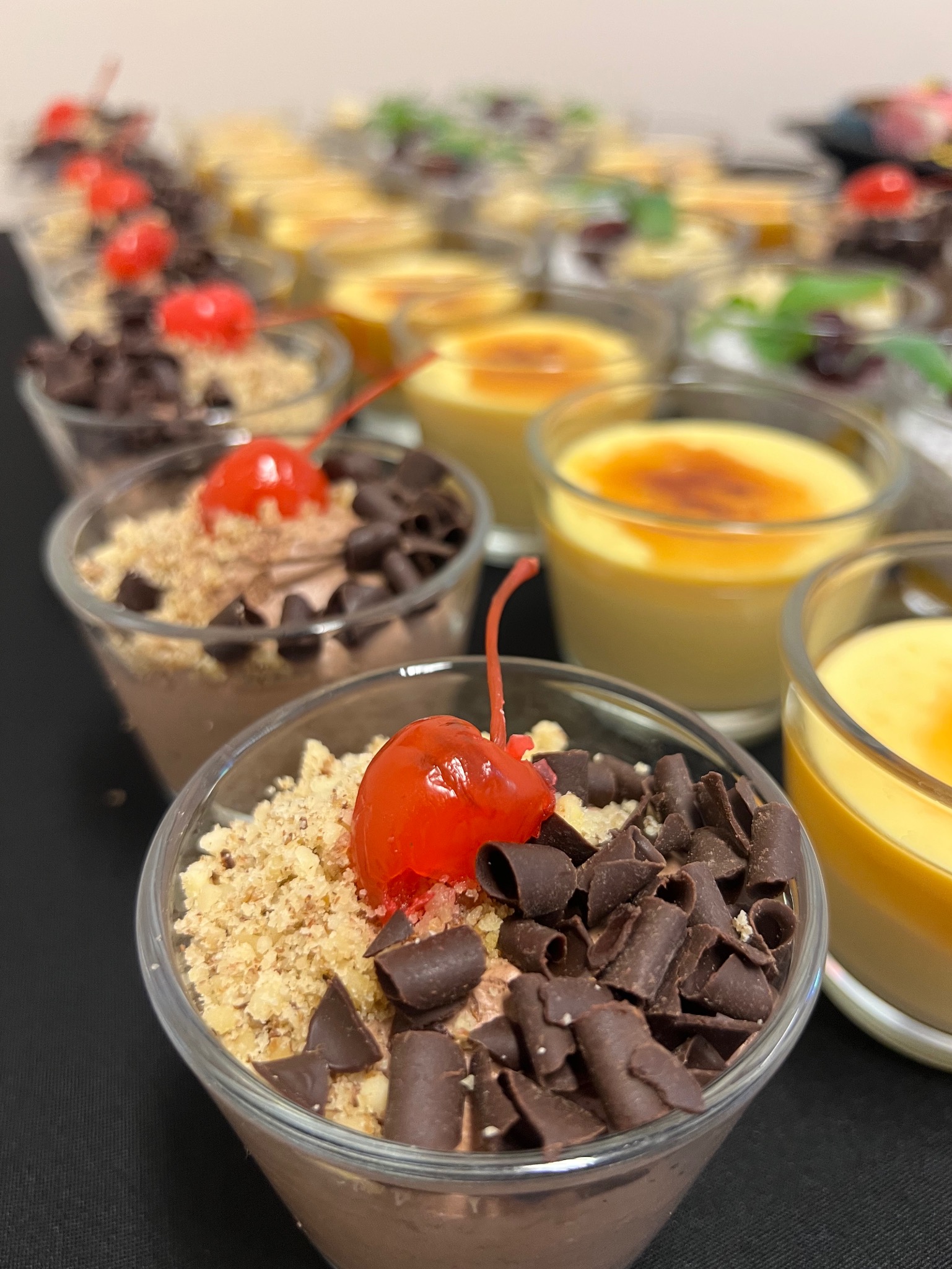 Deser w szklance: mus czekoladowy, kruszonka, wiśnia i płatki czekolady. W tle inne desery, w tym crème brûlée. Catering deserowy na ciemnym tle.