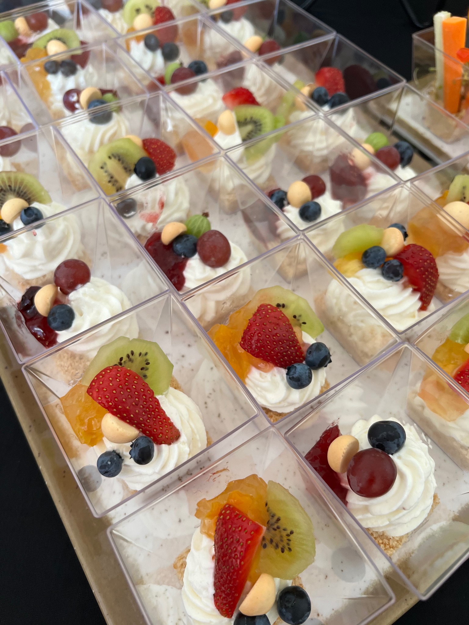 Deserowe mini porcje w przezroczystych pojemnikach: krem, owoce (kiwi, truskawki, winogrona, borówki), ułożone w rzędach na beżowej tacy, gotowe do serwowania.