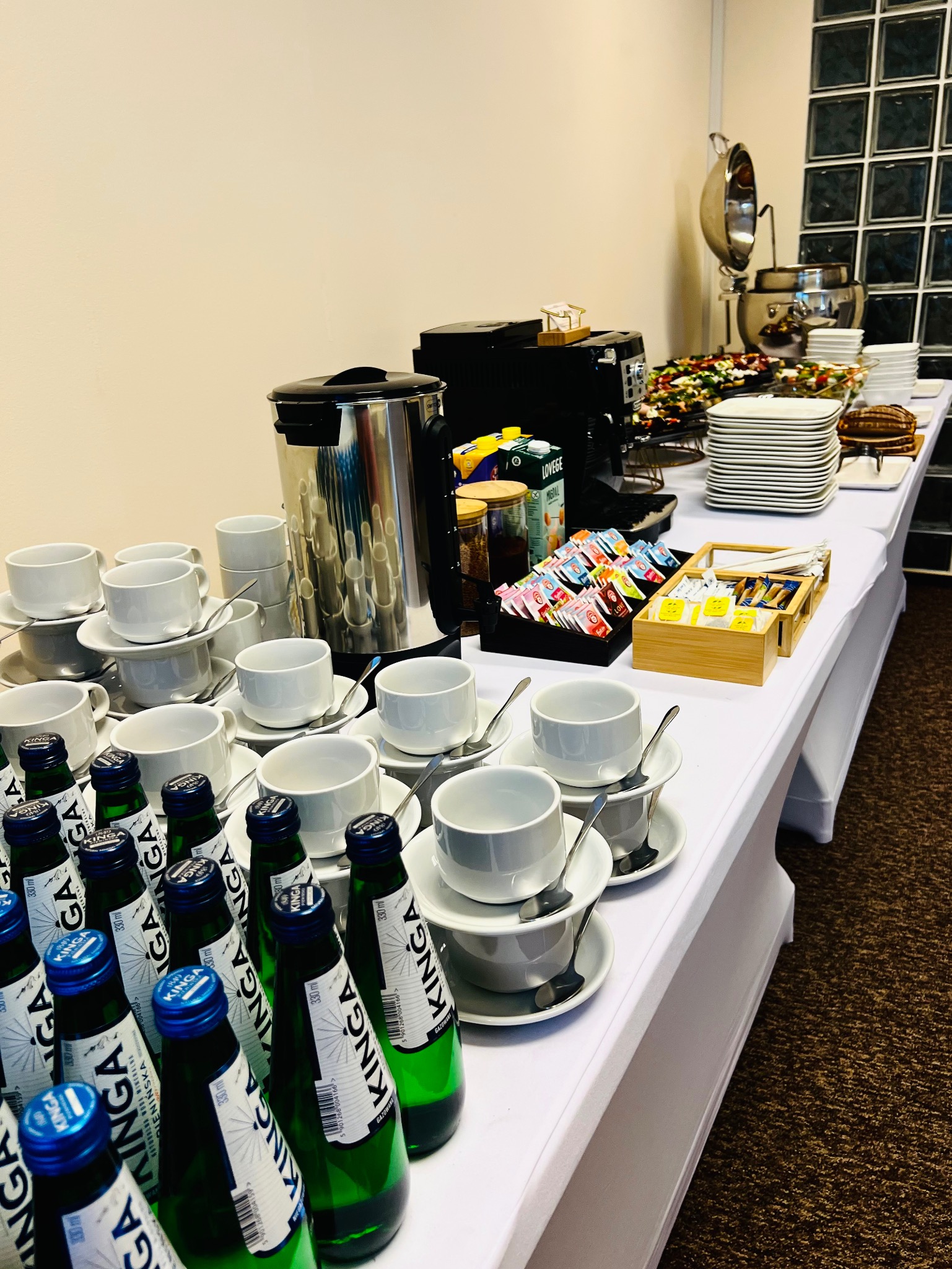 Stół cateringowy z wodą Kinga, kawą, herbatą, ciastami i sałatkami. Biały obrus, stosy filiżanek, ekspres do kawy. Idealny na firmowe wydarzenie.