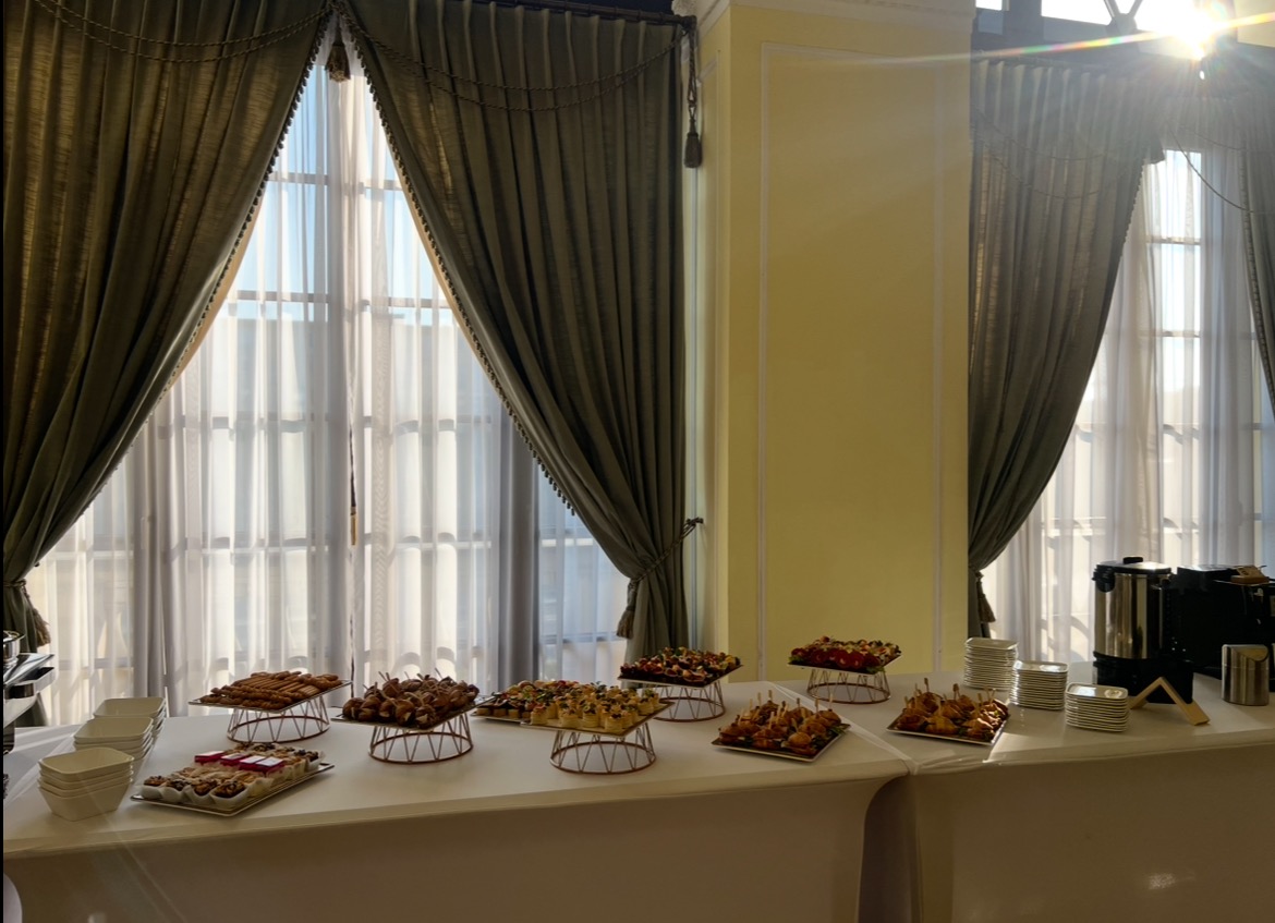 Elegancki stół cateringowy z przekąskami na paterach, tło stanowią okna z dekoracyjnymi zasłonami i jasne ściany. Widoczny ekspres do kawy.