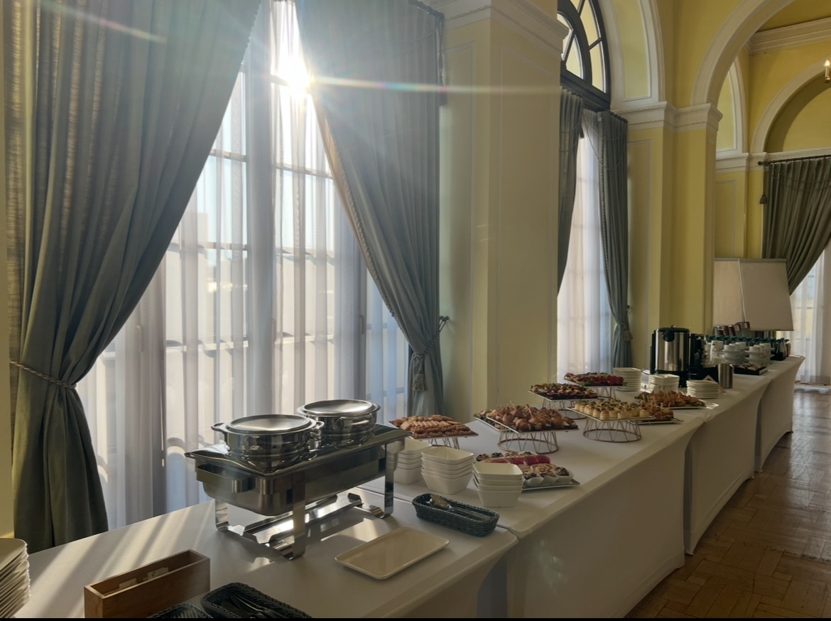 Elegancki stół cateringowy z bufetem potraw, ustawiony przy oknie z zasłonami w jasnym, przestronnym wnętrzu z kolumnami i łukami. Słońce wpada przez okno.