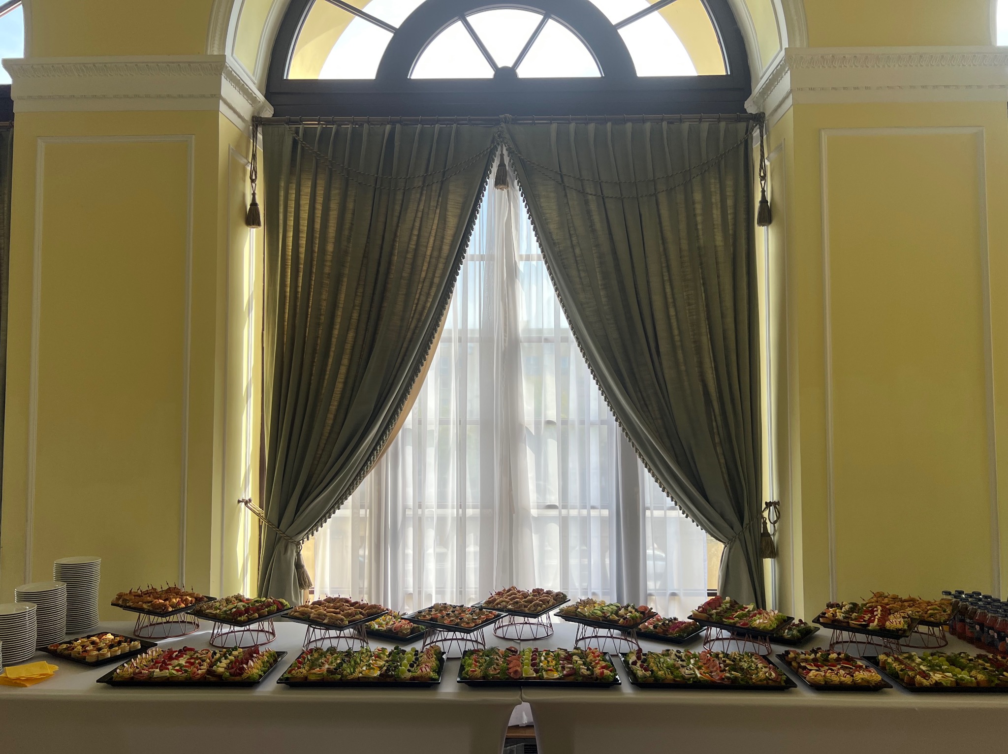 Stół cateringowy z przekąskami na czarnych tacach, ustawiony przed oknem z zasłonami w eleganckim wnętrzu z żółtymi ścianami i dekoracyjnymi detalami architektonicznymi.