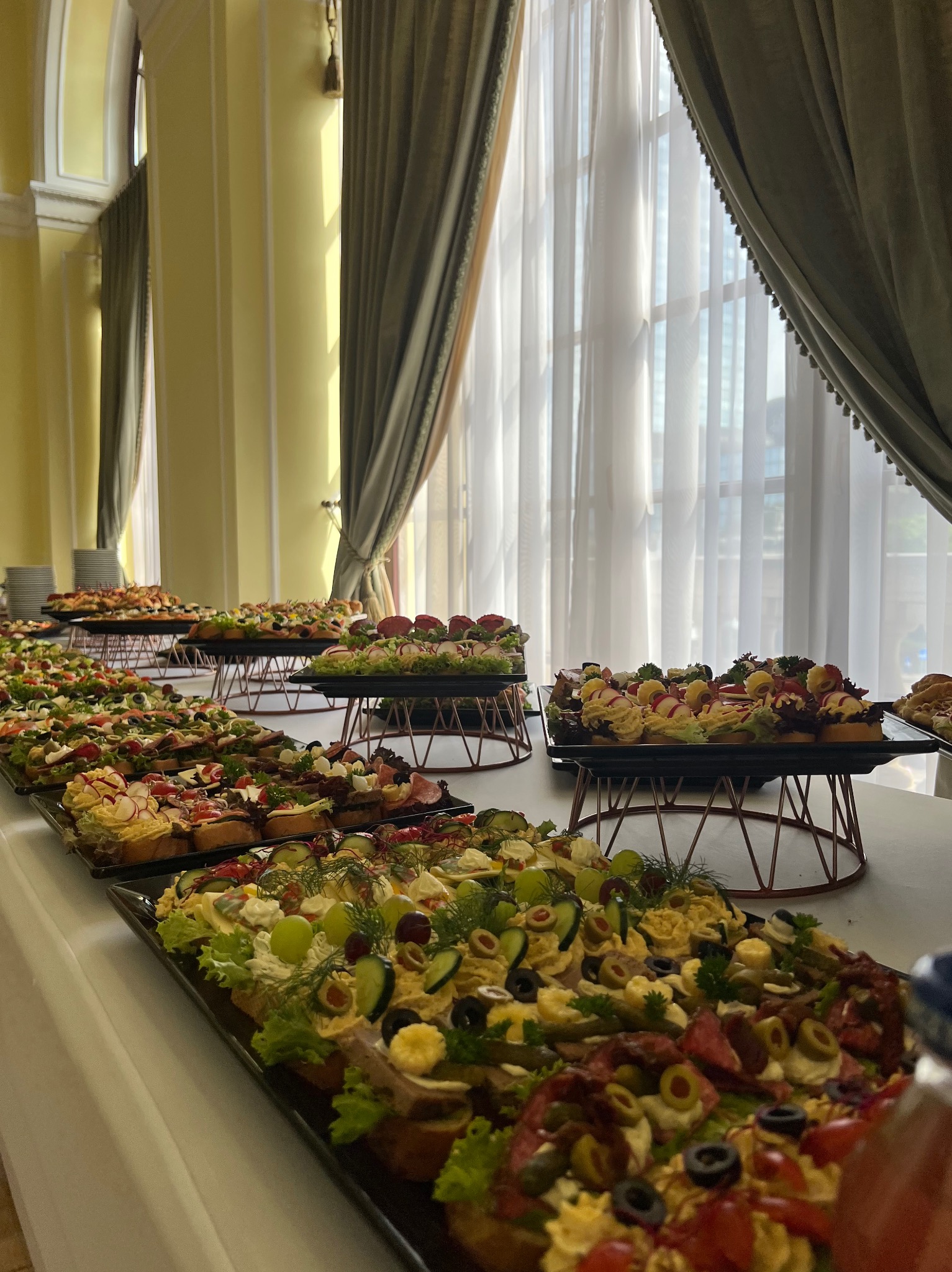 Stół cateringowy z kanapkami na ciemnych tacach, ustawiony w jasnym pomieszczeniu z kolumnami i zasłonami. Różnorodność składników i dekoracji.
