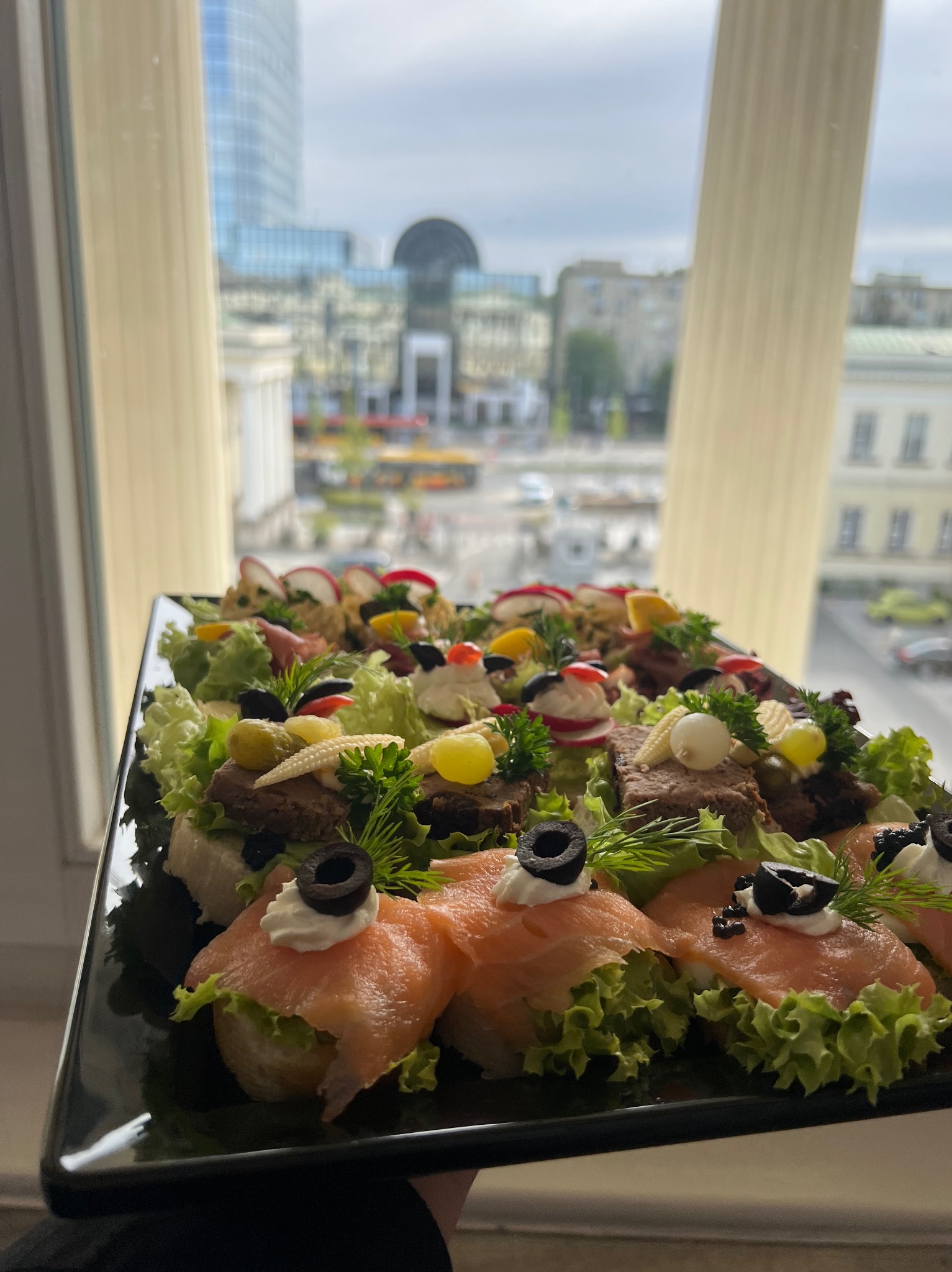 Elegancka taca z kanapkami bankietowymi, dekorowanymi sałatą, łososiem, oliwkami i ziołami, na tle okna z widokiem na miasto.
