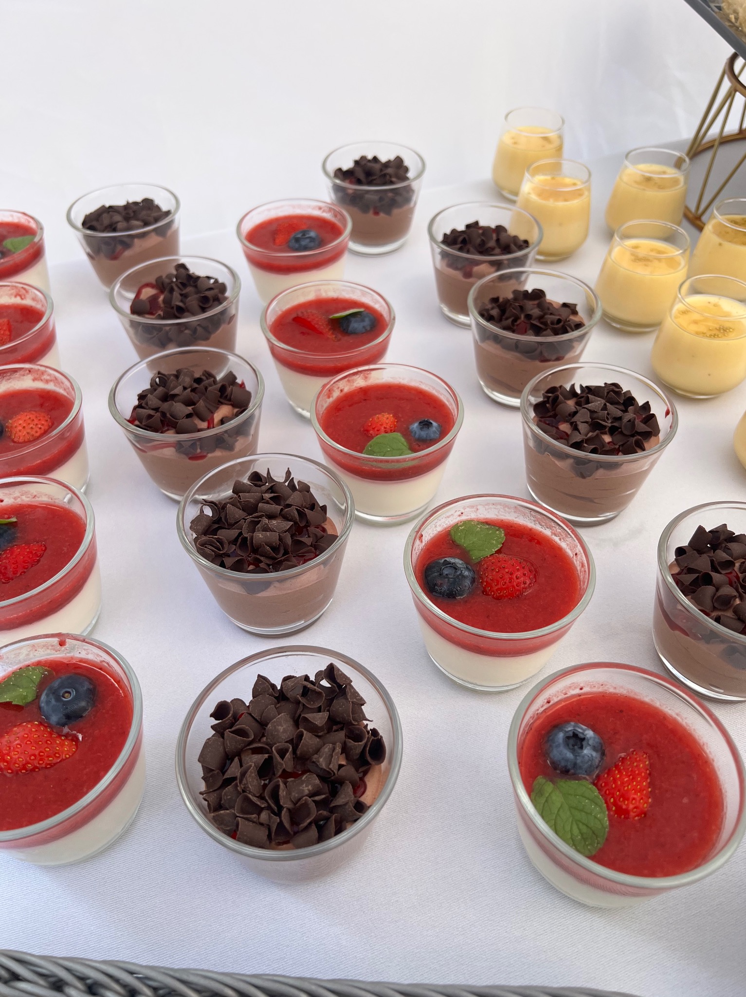 Deserki w szklanych pucharkach: panna cotta z sosem truskawkowym i owocami, mus czekoladowy z wiórkami. Elegancki catering na białym obrusie.