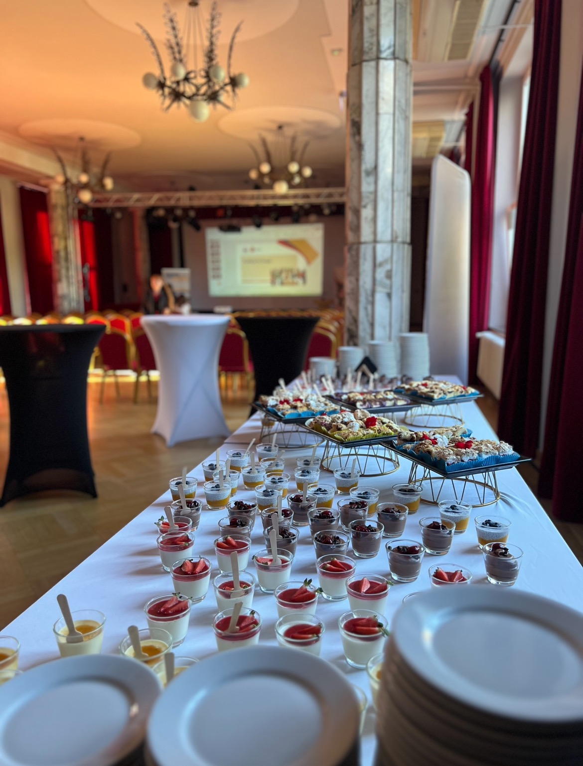 Stół cateringowy z deserami w szklankach i przekąskami na paterach, przygotowany na eleganckie wydarzenie w sali z kolumnami i sceną.