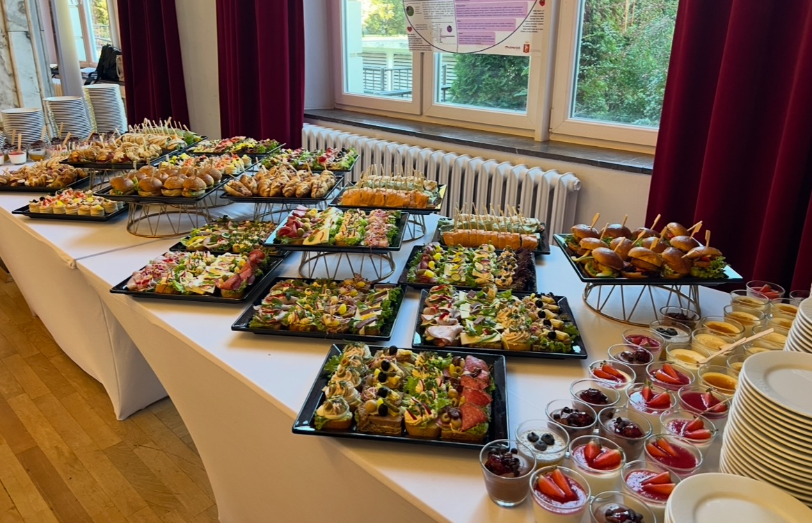 Stół cateringowy z różnorodnymi przekąskami: mini burgery, kanapki, sałatki i desery w szklankach. Biały obrus, eleganckie podesty, talerze w stosach. Ujęcie z boku.