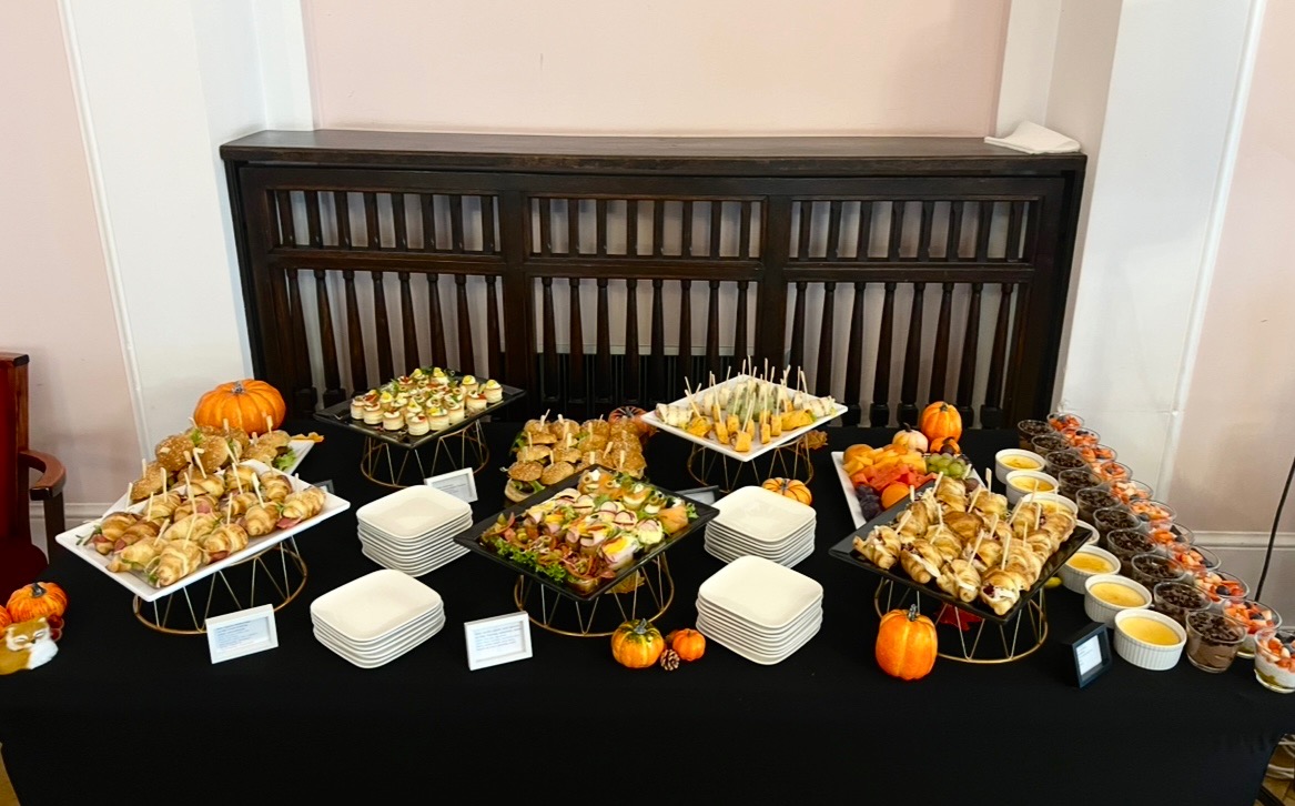 Jesienny bufet cateringowy: różnorodne przekąski na eleganckich stojakach, dynie jako dekoracja, czarny obrus, białe ściany. Widoczne mini burgery, roladki, desery w szklankach.