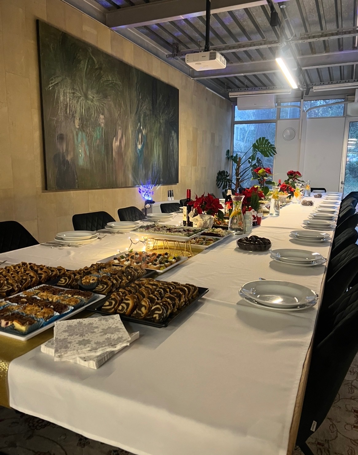 Długi stół z białym obrusem, zastawiony talerzami i przekąskami. W tle obraz na ścianie i projektor pod sufitem. Dekoracje świąteczne i oświetlenie tworzą przytulną atmosferę.