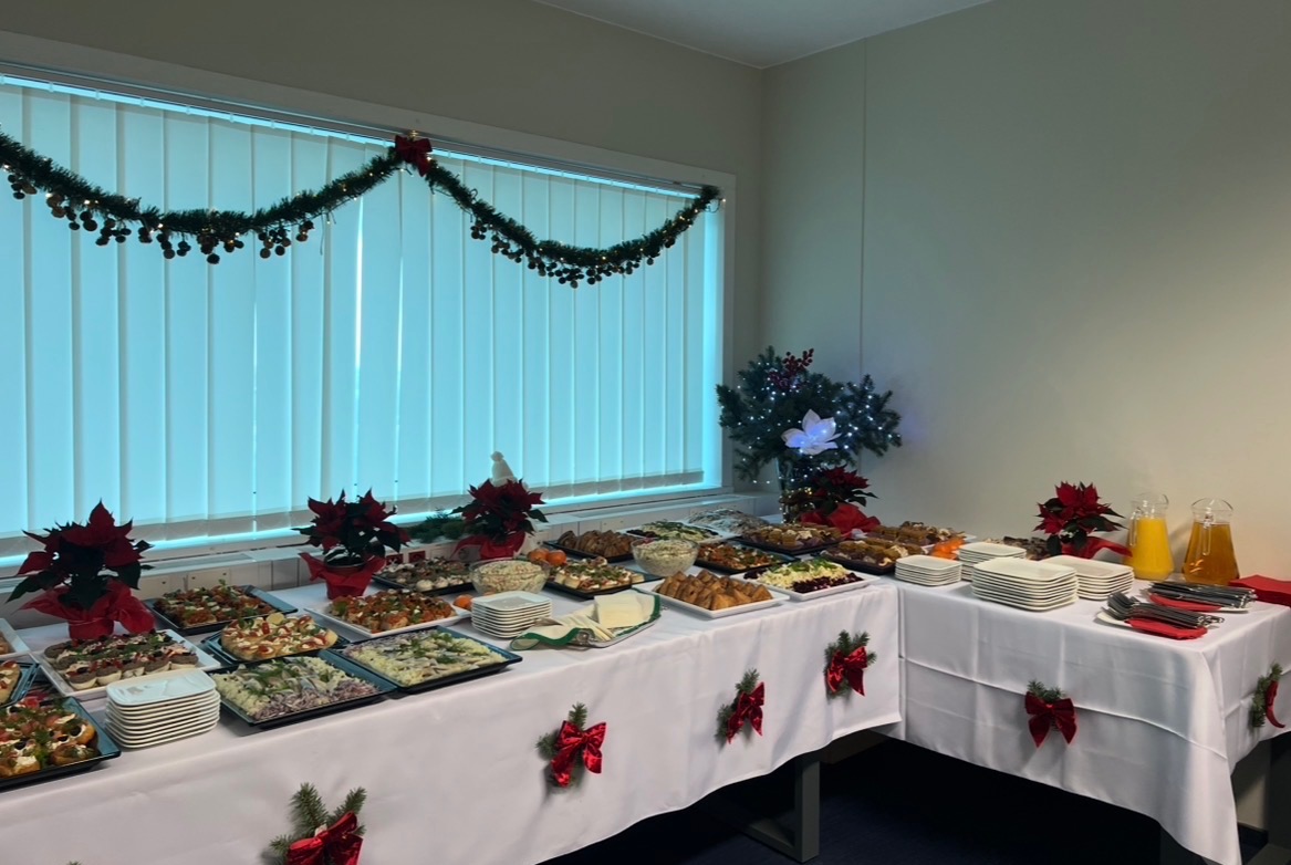 Świąteczny stół cateringowy z dekoracjami: stroiki z poinsecji, girlanda nad oknem, różnorodne dania, napoje w dzbankach. Biały obrus z czerwonymi kokardami.
