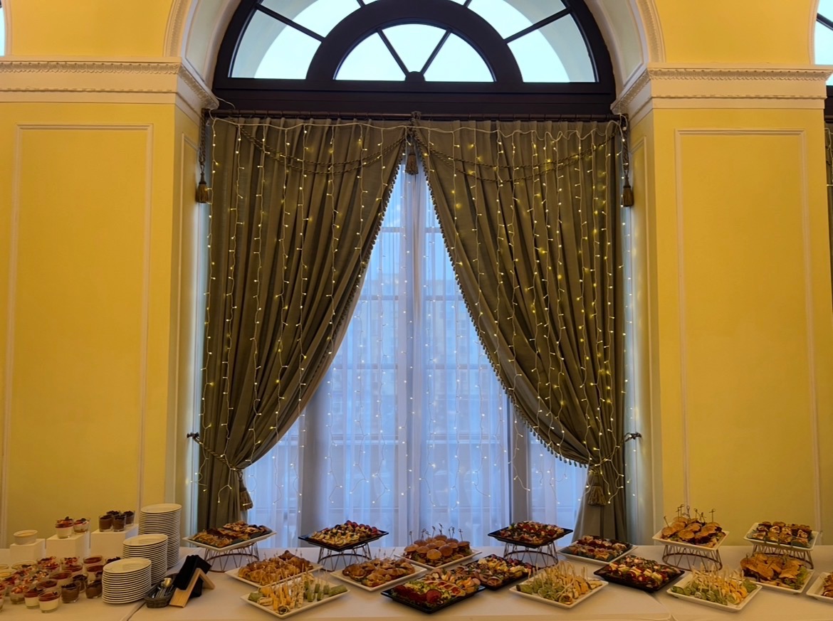 Elegancki stół cateringowy z przekąskami na tle okna z dekoracyjnymi zasłonami i lampkami, w jasnym wnętrzu. Różnorodność kanapek, deserów i owoców.
