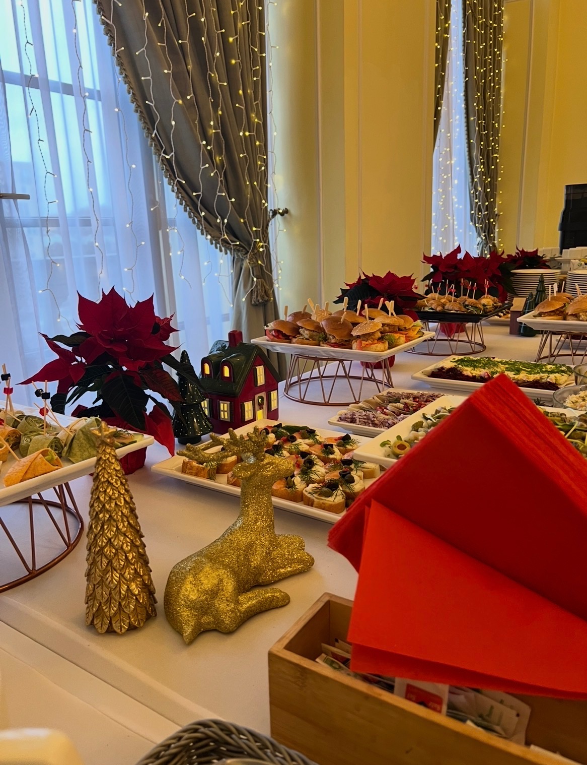 Świąteczny stół cateringowy z dekoracjami: złoty renifer, choinka, poinsecje i różnorodne przekąski na białych paterach. Okno z girlandami w tle.