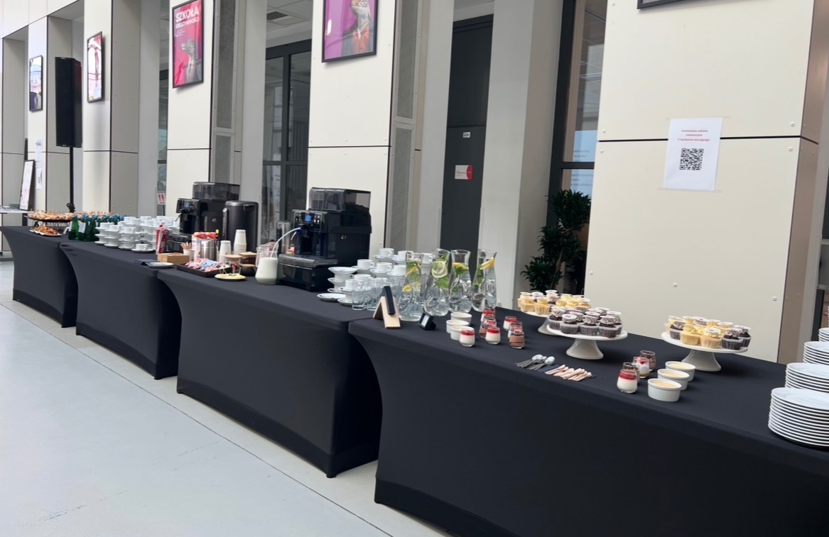 Stół cateringowy z czarnym obrusem: ekspresy do kawy, napoje w dzbankach, babeczki na paterach, desery w słoiczkach. W tle jasne wnętrze z kolumnami i plakatami.