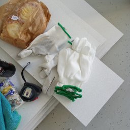 Zakład Ogólnobudowlany Michał łakomy - Stos białych płyt izolacyjnych leżących na szarym podłożu, obok rękawice robocze z zielonymi ściągaczami, zwinięta miarka, paczka chusteczek, napój energetyczny i ciastka w papierowej torbie.