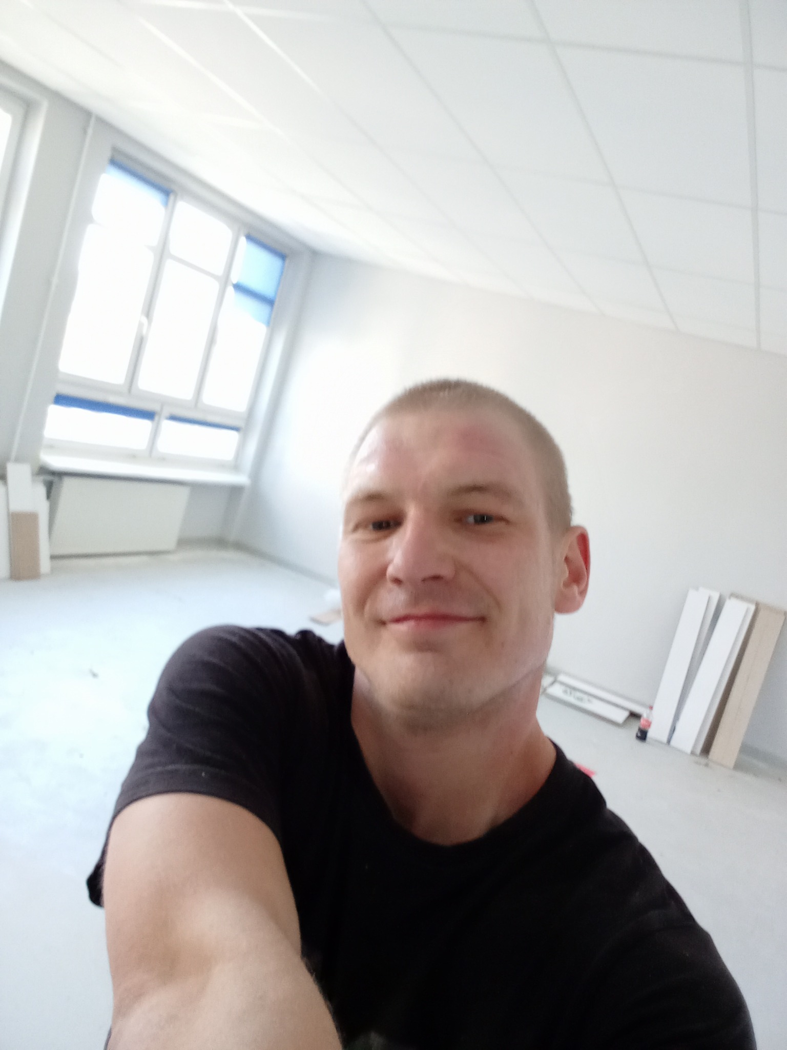 Mężczyzna robi selfie w pustym, jasnym pokoju z oknami dachowymi i panelami pod ścianą, prawdopodobnie po remoncie.