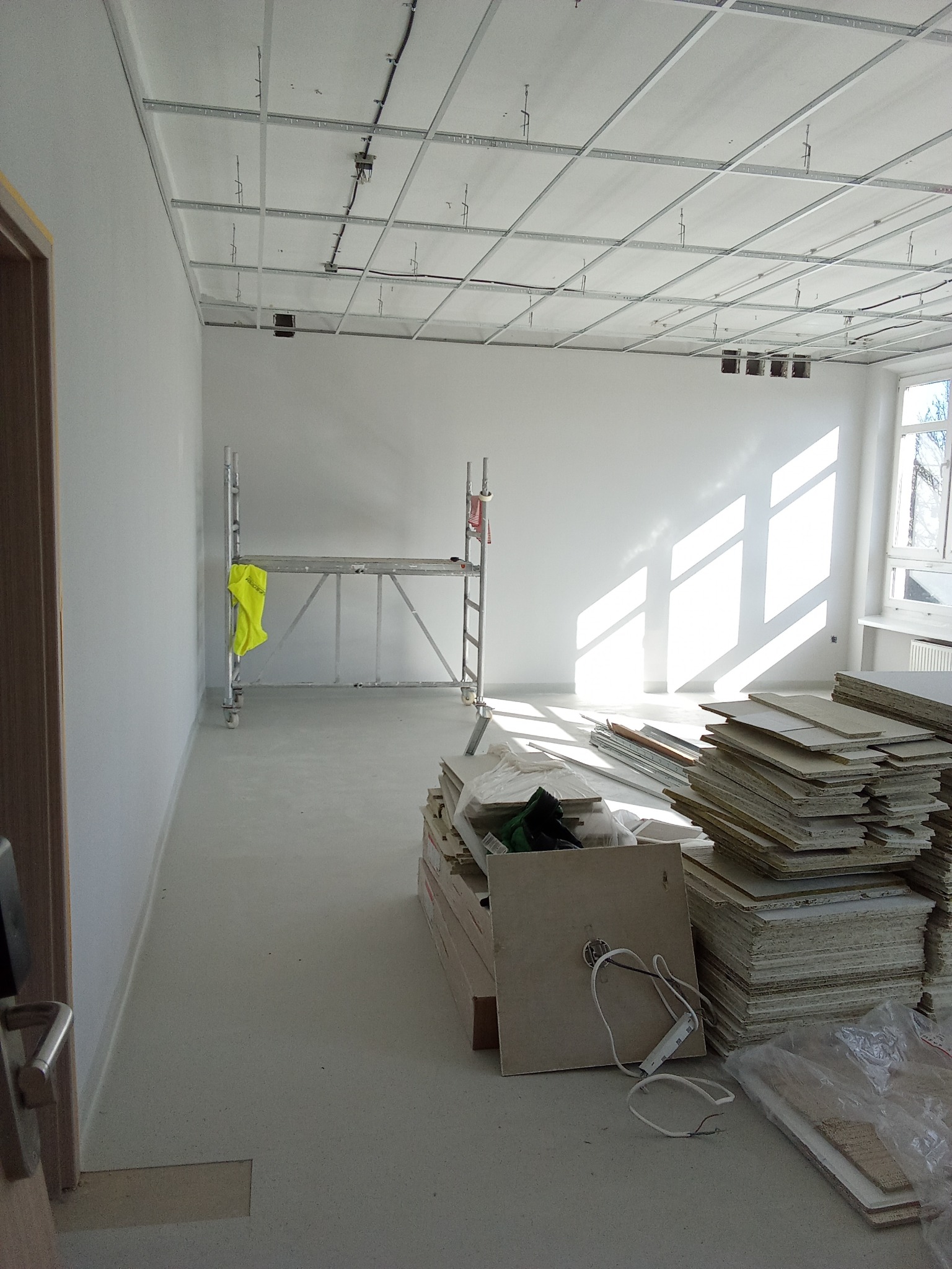 Wnętrze w trakcie prac wykończeniowych: widoczne rusztowanie, stosy płyt, przygotowane do montażu podwieszanego sufitu w jasnym pomieszczeniu z oknem.