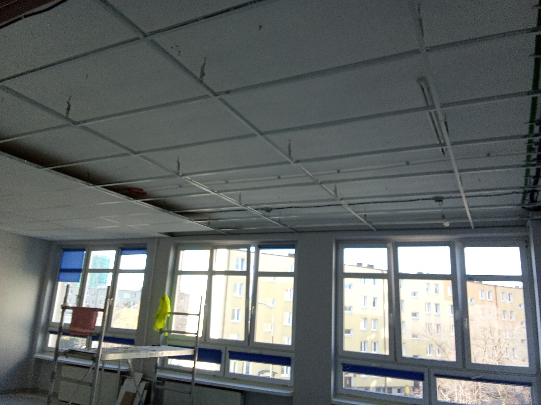 Montaż sufitu podwieszanego z widoczną konstrukcją metalową i częściowo ułożonymi panelami w pomieszczeniu z oknami i rusztowaniem.