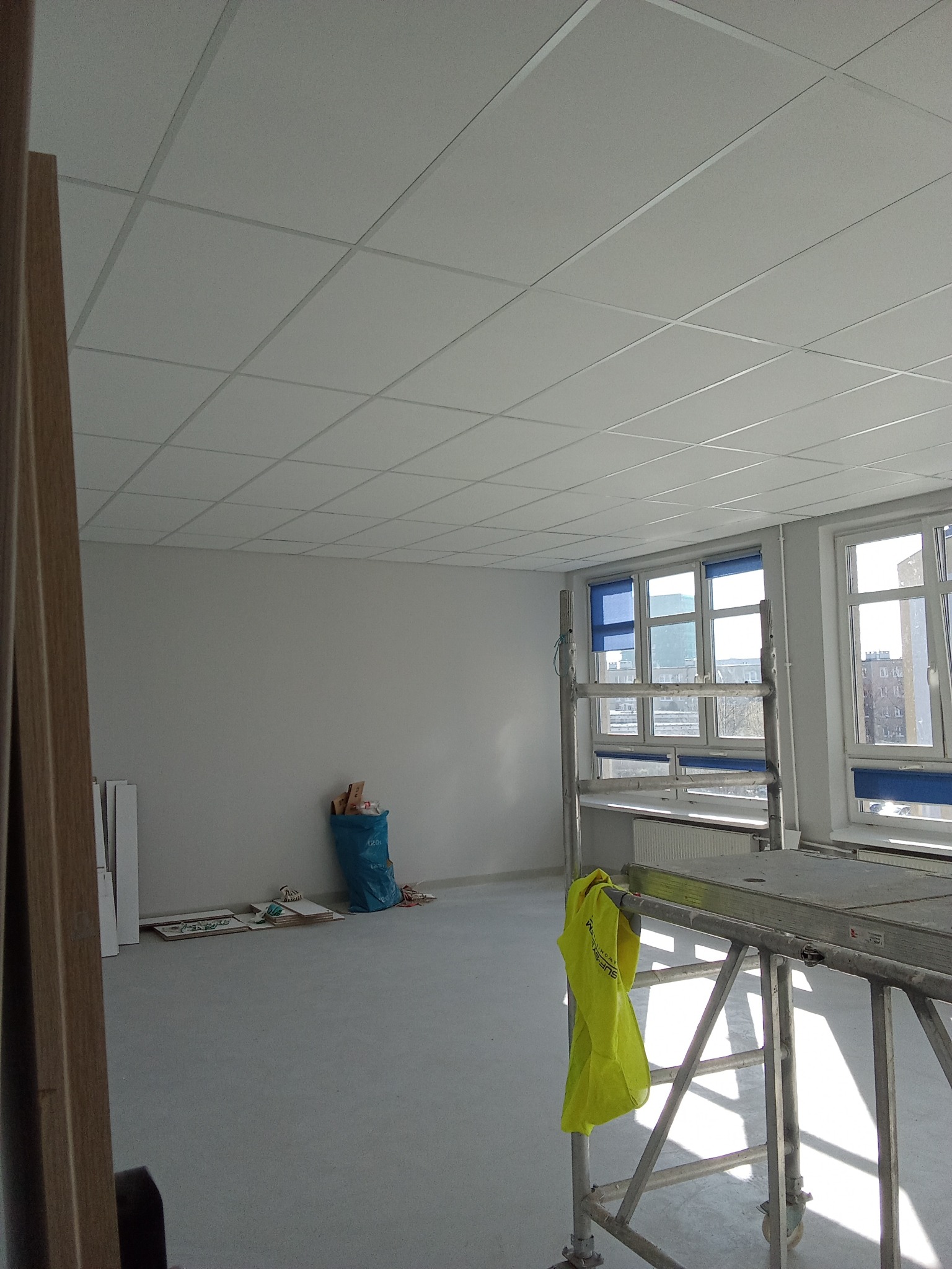Wnętrze pokoju w trakcie remontu, widoczne rusztowanie z jaskrawożółtą kamizelką, ściany przygotowane do malowania, sufit podwieszany kasetonowy, okna z niebieskimi roletami, worek z odpadami...