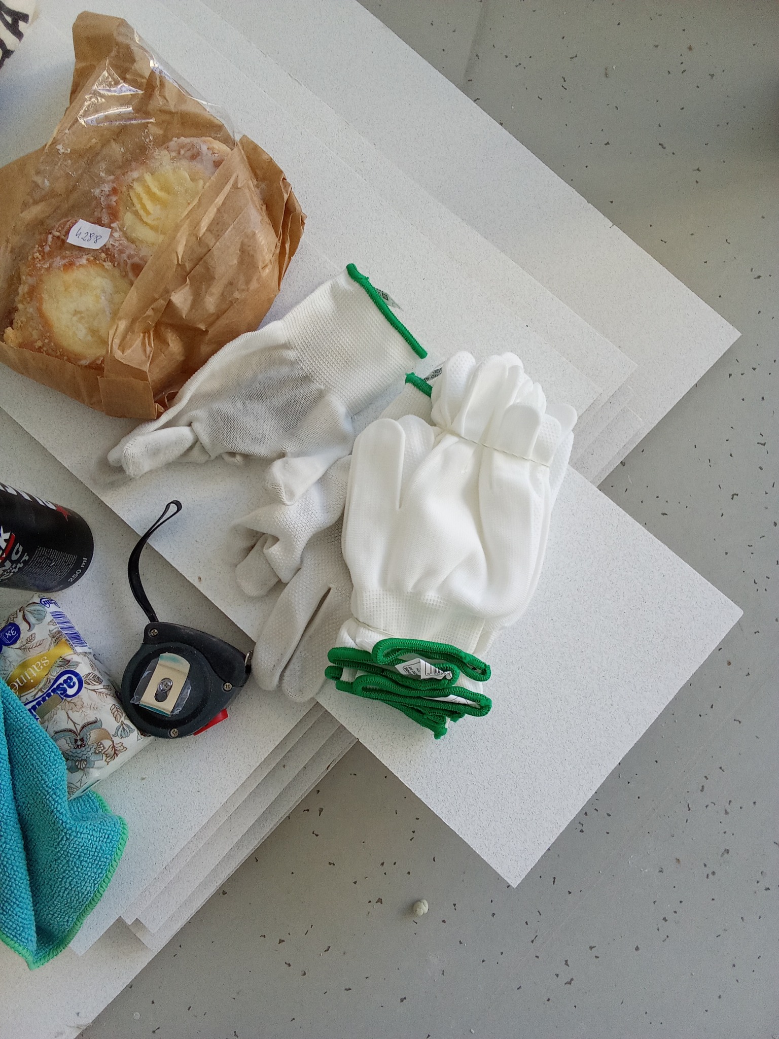 Stos białych płyt izolacyjnych leżących na szarym podłożu, obok rękawice robocze z zielonymi ściągaczami, zwinięta miarka, paczka chusteczek, napój energetyczny i ciastka w papierowej torbie.