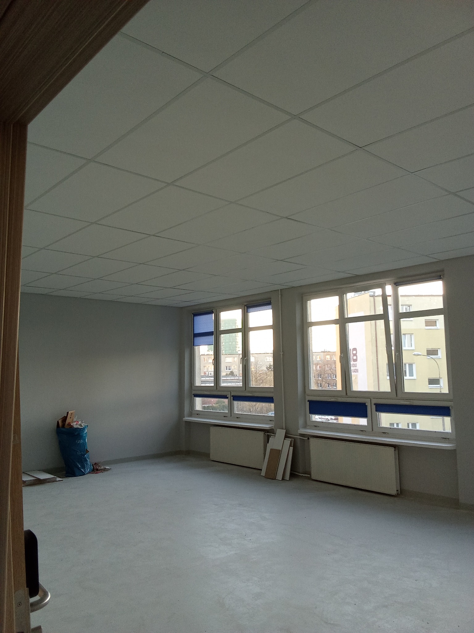 Puste, szare pomieszczenie po remoncie z trzema oknami z niebieskimi roletami, grzejnikami pod oknami, workiem śmieci w rogu i kafelkowym sufitem.