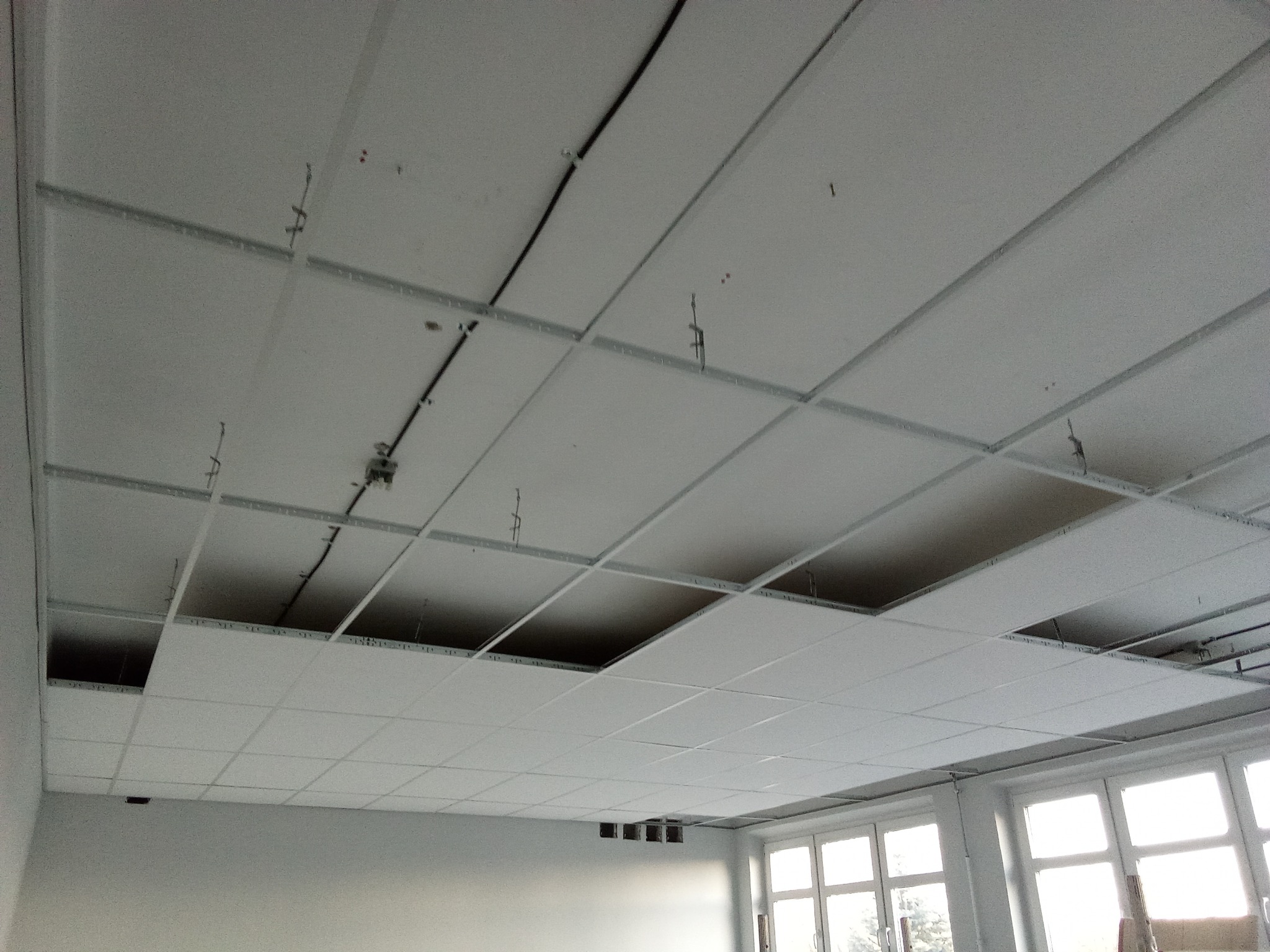 Montaż sufitu podwieszanego kasetonowego z widocznymi elementami konstrukcyjnymi i instalacją elektryczną, kilka kasetonów brakuje.