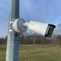 HANSSA Andrzej Kaczmarek - Kamera monitoringu Dahua zamontowana na metalowym słupie, widoczne mocowania i fragment krajobrazu z drzewami w tle.
