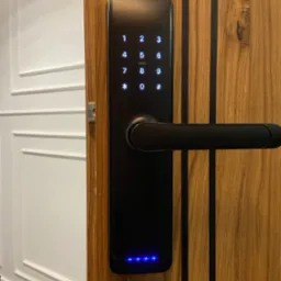 Elegancki, czarny wideodomofon z klawiaturą numeryczną i czytnikiem kart zamontowany na drewnianych drzwiach w Aleksandrowie Łódzkim, podświetlony niebieskimi diodami.