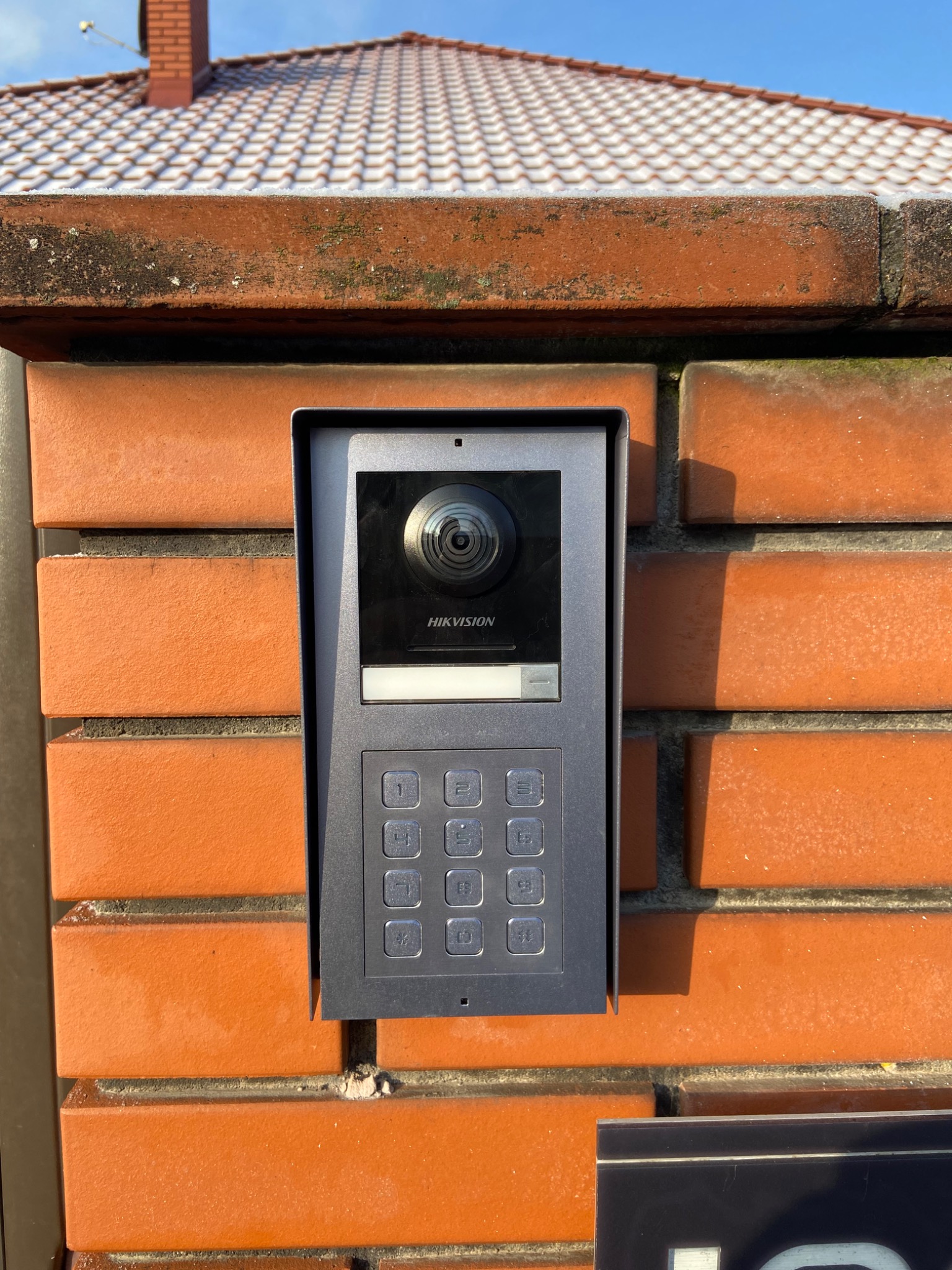 Zewnętrzny panel wideodomofonu Hikvision zamontowany na ceglanym murze z widocznym dachem pokrytym śniegiem w tle.