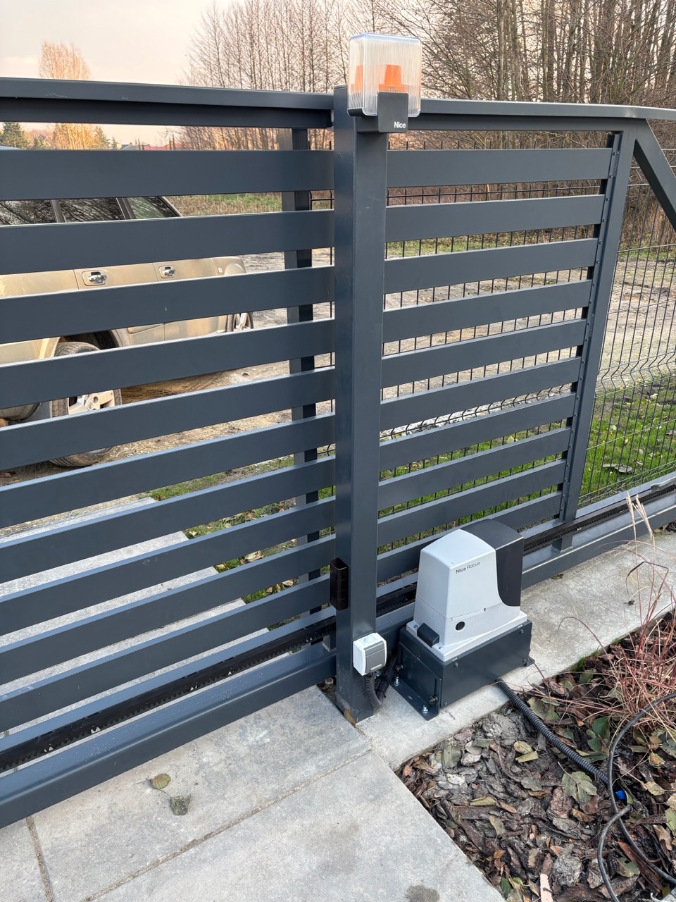 Szara, przesuwna brama wjazdowa z automatyką Nice Robus, lampą ostrzegawczą i listwą zębatą, zamontowana na betonowej kostce.