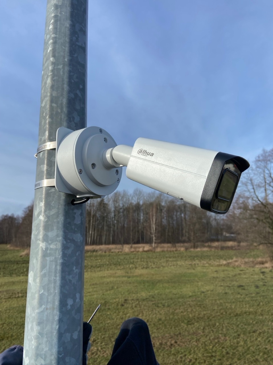 Kamera monitoringu Dahua zamontowana na metalowym słupie, widoczne mocowania i fragment krajobrazu z drzewami w tle.