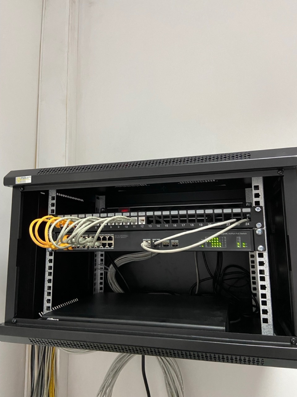 Czarny panel krosowniczy z podłączonymi kablami Ethernet w szafie rack, widoczne porty 1-24, część kabli w kolorze żółtym i białym, diody sygnalizacyjne aktywne.