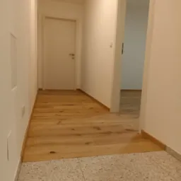 Jasne panele podłogowe w korytarzu, łączące się z terakotą. Widoczne białe ściany i drzwi, minimalistyczny styl wykończenia wnętrza. Oświetlenie ciepłe, naturalne.