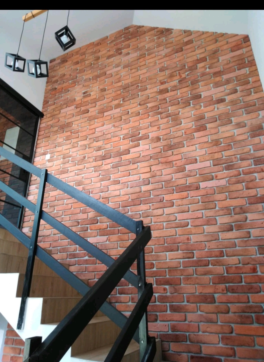 Ściana z cegły klinkierowej wewnątrz domu, widoczne schody z czarną balustradą i nowoczesne, geometryczne lampy wiszące.