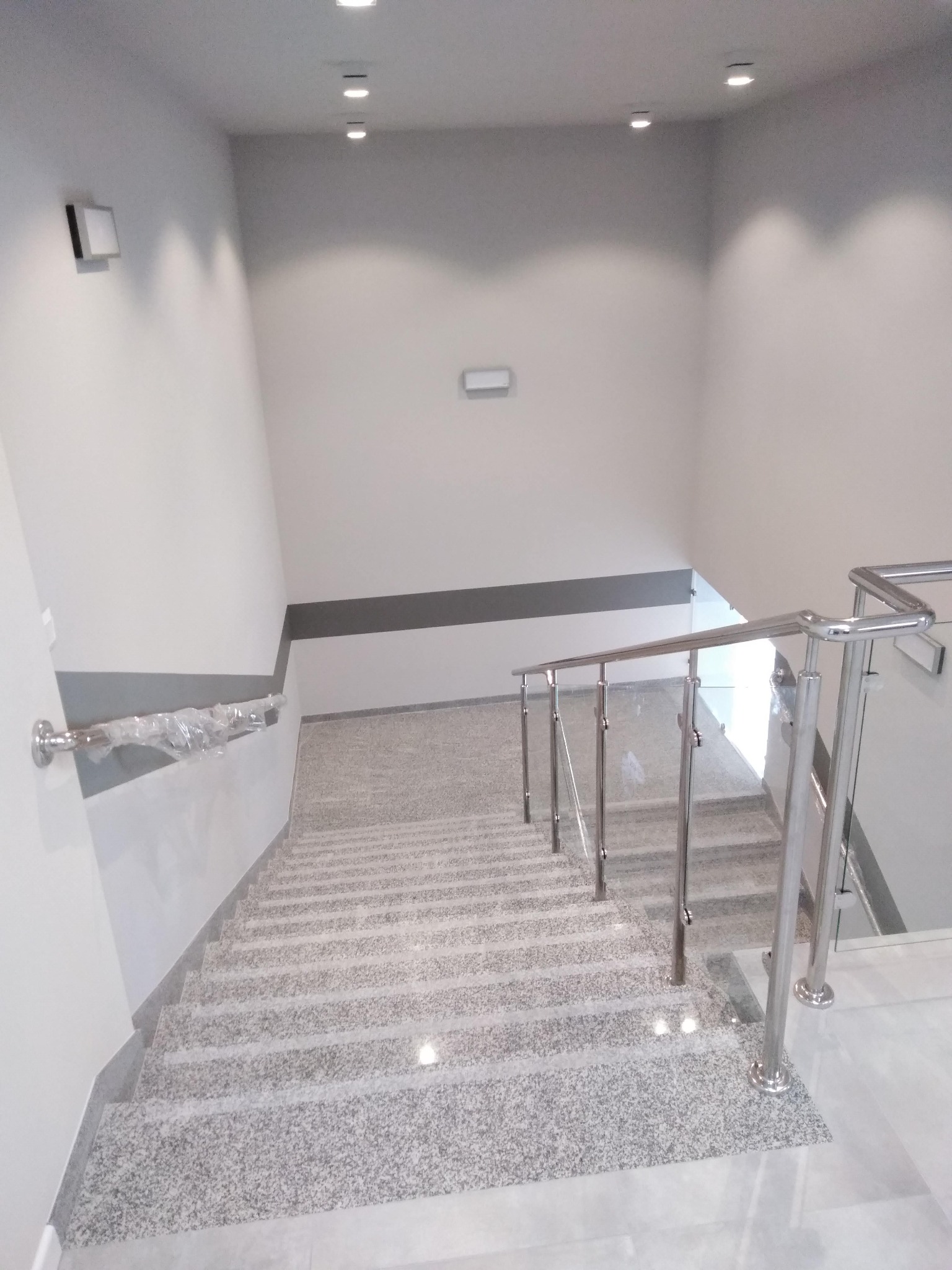 Klatka schodowa po remoncie. Szare, granitowe schody z metalową balustradą i szklanymi panelami. Ściany pomalowane na biało z szarym pasem. Widoczne oświetlenie.