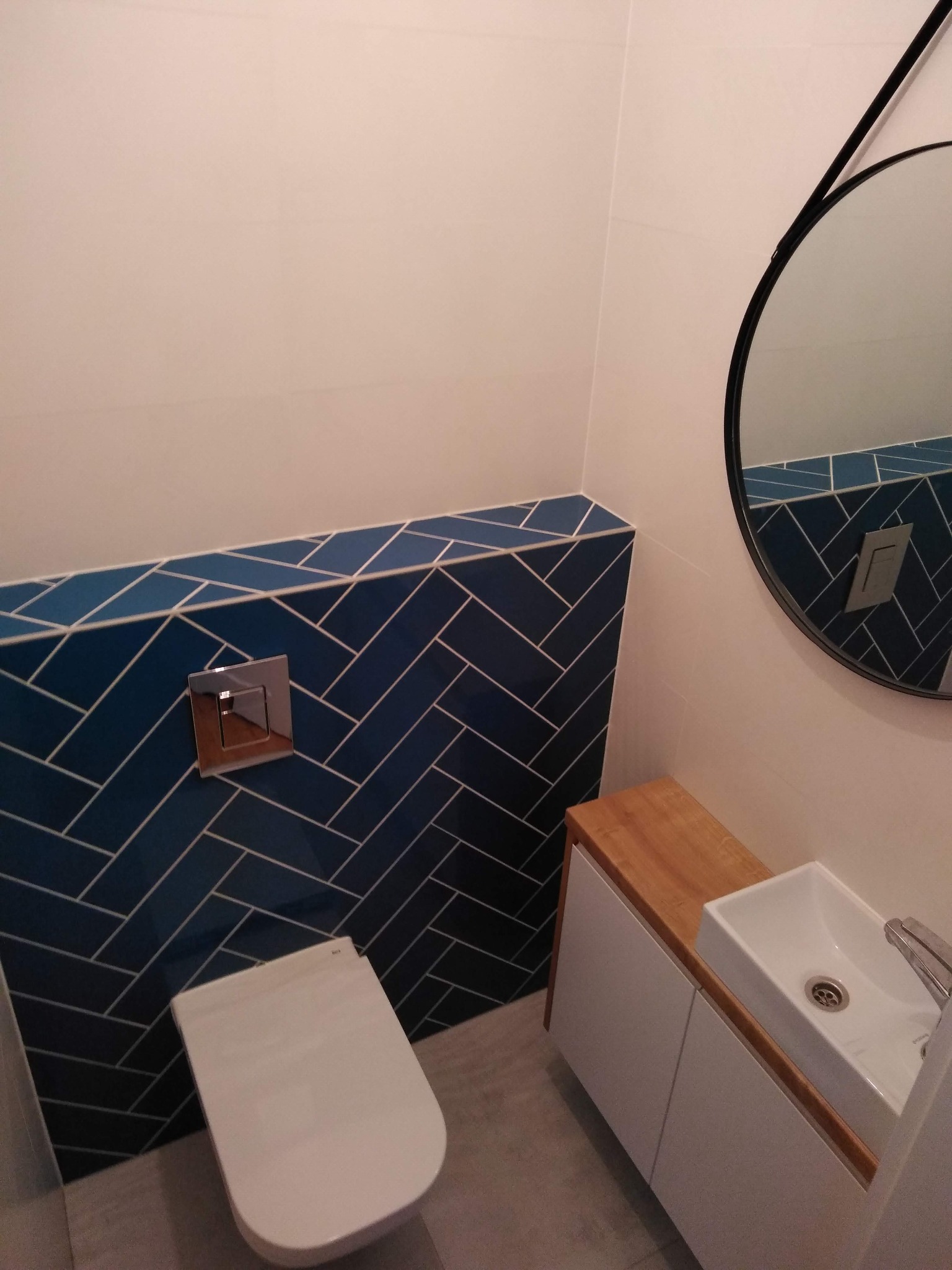 Wykończona łazienka: wisząca toaleta, umywalka na szafce z drewnianym blatem i lustro w czarnej ramie. Na ścianie ciemnoniebieskie płytki ułożone w jodełkę.