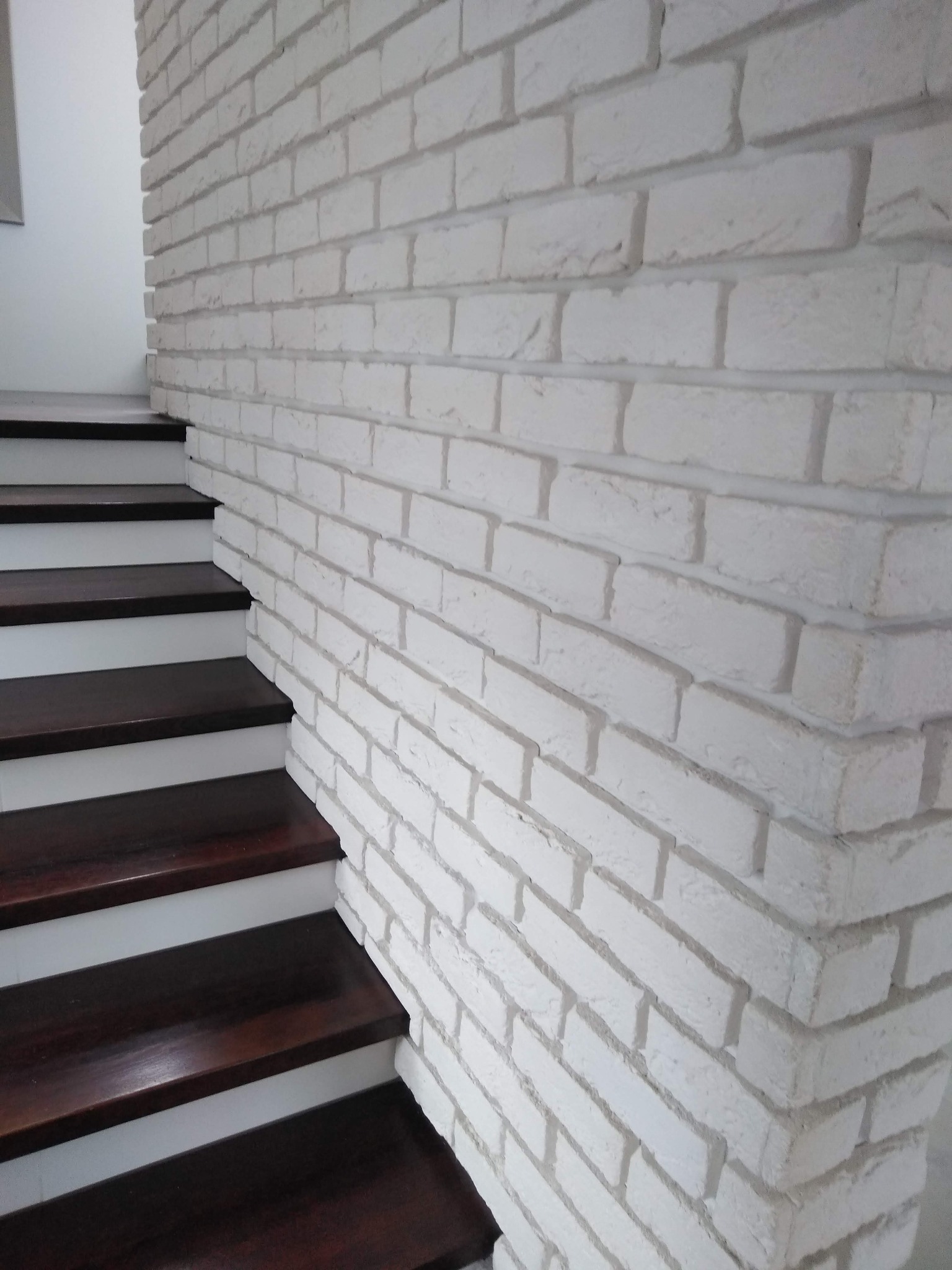 Biała ściana z cegły dekoracyjnej sąsiadująca z ciemnymi, drewnianymi schodami. Minimalistyczne wykończenie wnętrza, kontrast materiałów.
