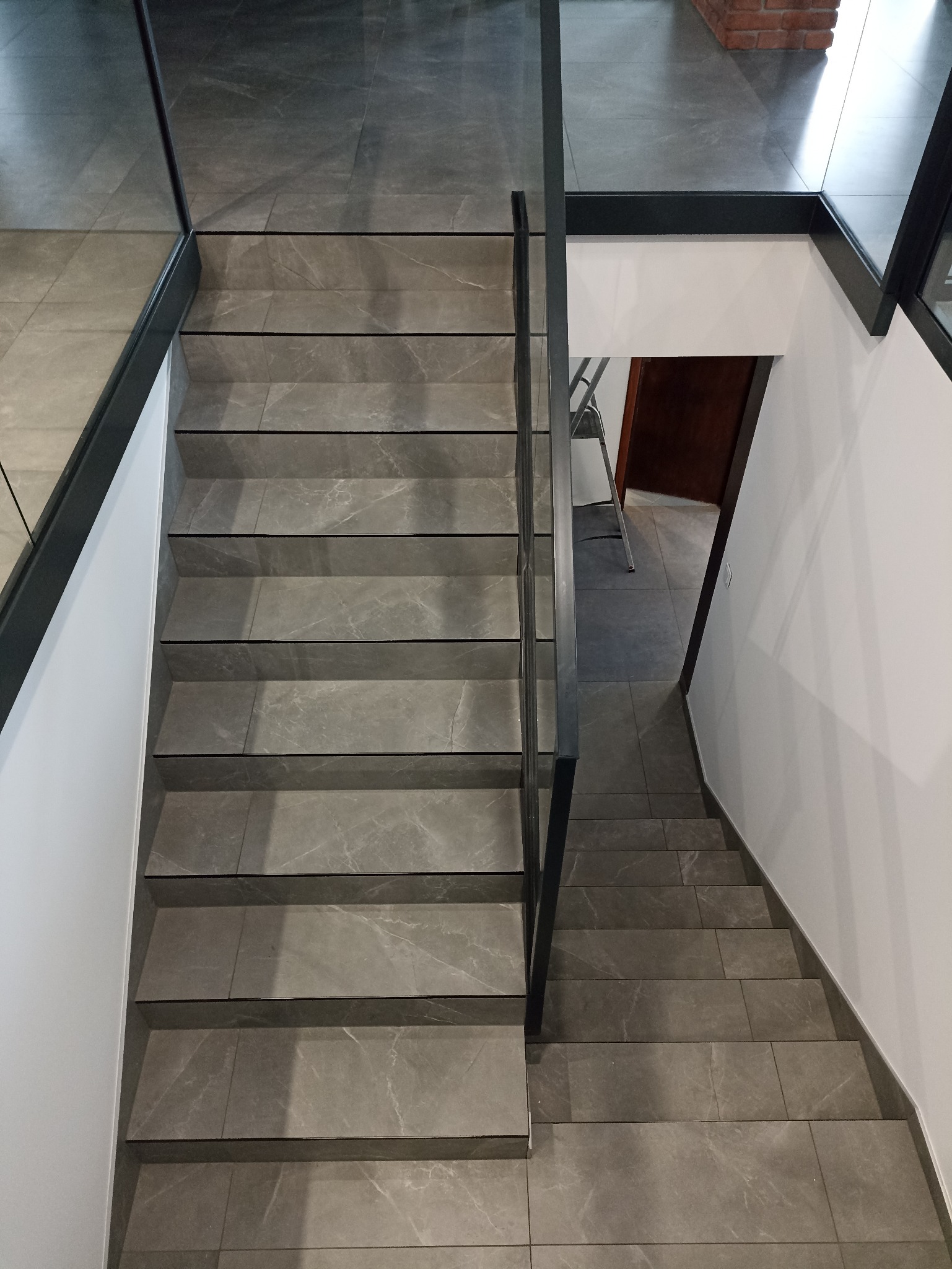 Wykończone schody z szarych płytek imitujących marmur, z czarnymi listwami i szklaną balustradą, widziane z góry. W tle widoczny fragment ściany z cegły.