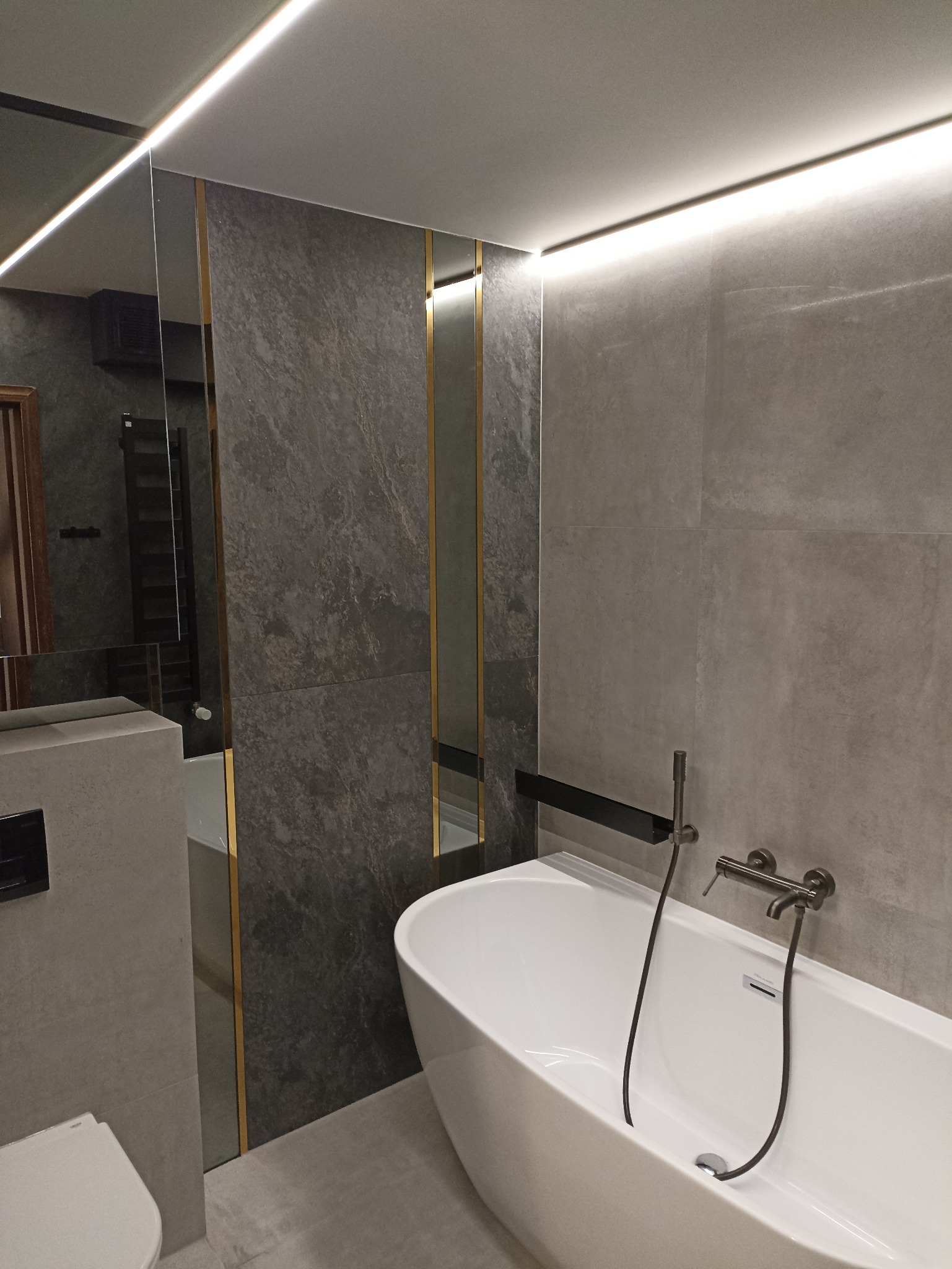 Nowoczesna łazienka z wolnostojącą wanną i szarą płytką imitującą kamień, oświetlona ledowym światłem. Lustro z podświetleniem i złotymi akcentami.