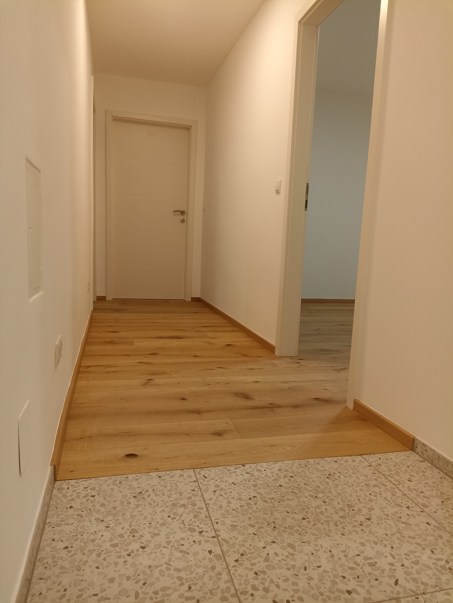 Jasne panele podłogowe w korytarzu, łączące się z terakotą. Widoczne białe ściany i drzwi, minimalistyczny styl wykończenia wnętrza. Oświetlenie ciepłe, naturalne.