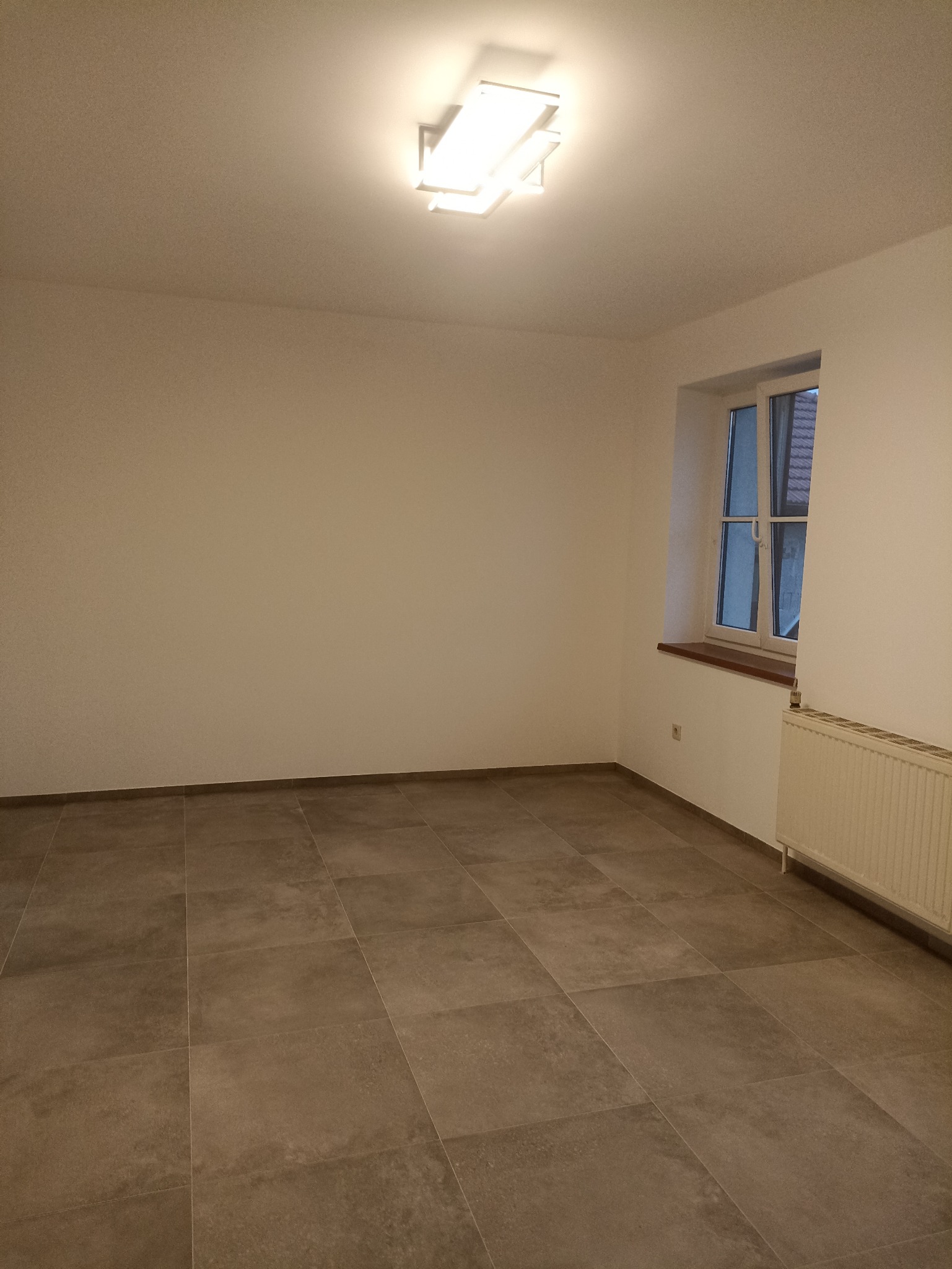 Wnętrze po remoncie: świeżo pomalowane ściany w odcieniach bieli, brązowe płytki podłogowe, okno z drewnianym parapetem i biały grzejnik. Nowoczesna lampa sufitowa.