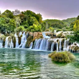 Kaskadowy wodospad z zielonymi brzegami i bujną roślinnością, woda o szmaragdowym odcieniu, pochmurne niebo w tle.