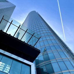 BMC Foto - Wysoki, nowoczesny biurowiec z zaokrągloną fasadą odbijający inne budynki, widziany z dołu na tle błękitnego nieba z śladem kondensacyjnym samolotu.