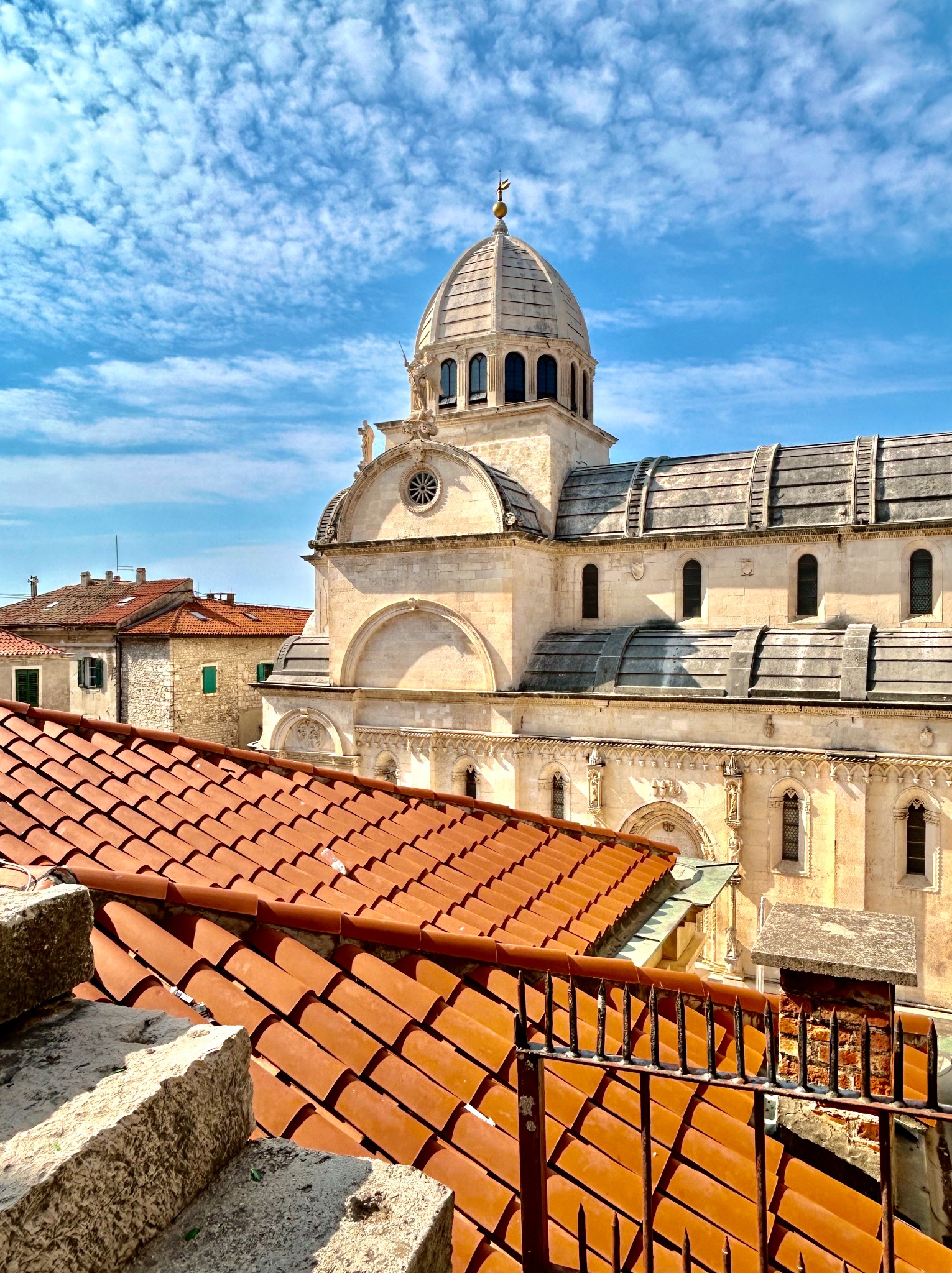 Widok z góry na katedrę z kopułą i czerwonymi dachówkami w Szybeniku, Chorwacja, przy błękitnym niebie z chmurami.