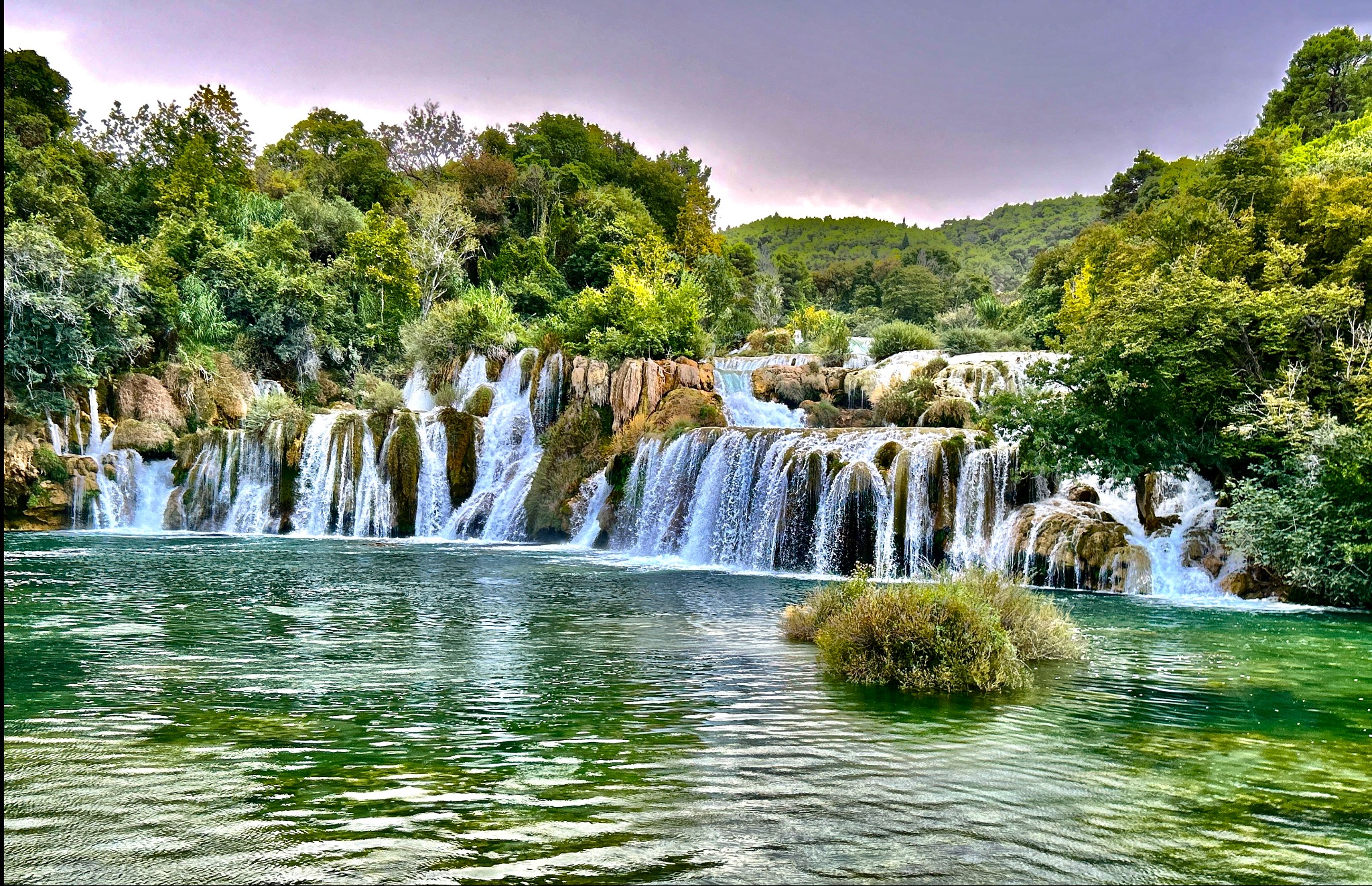 Kaskadowy wodospad z zielonymi brzegami i bujną roślinnością, woda o szmaragdowym odcieniu, pochmurne niebo w tle.