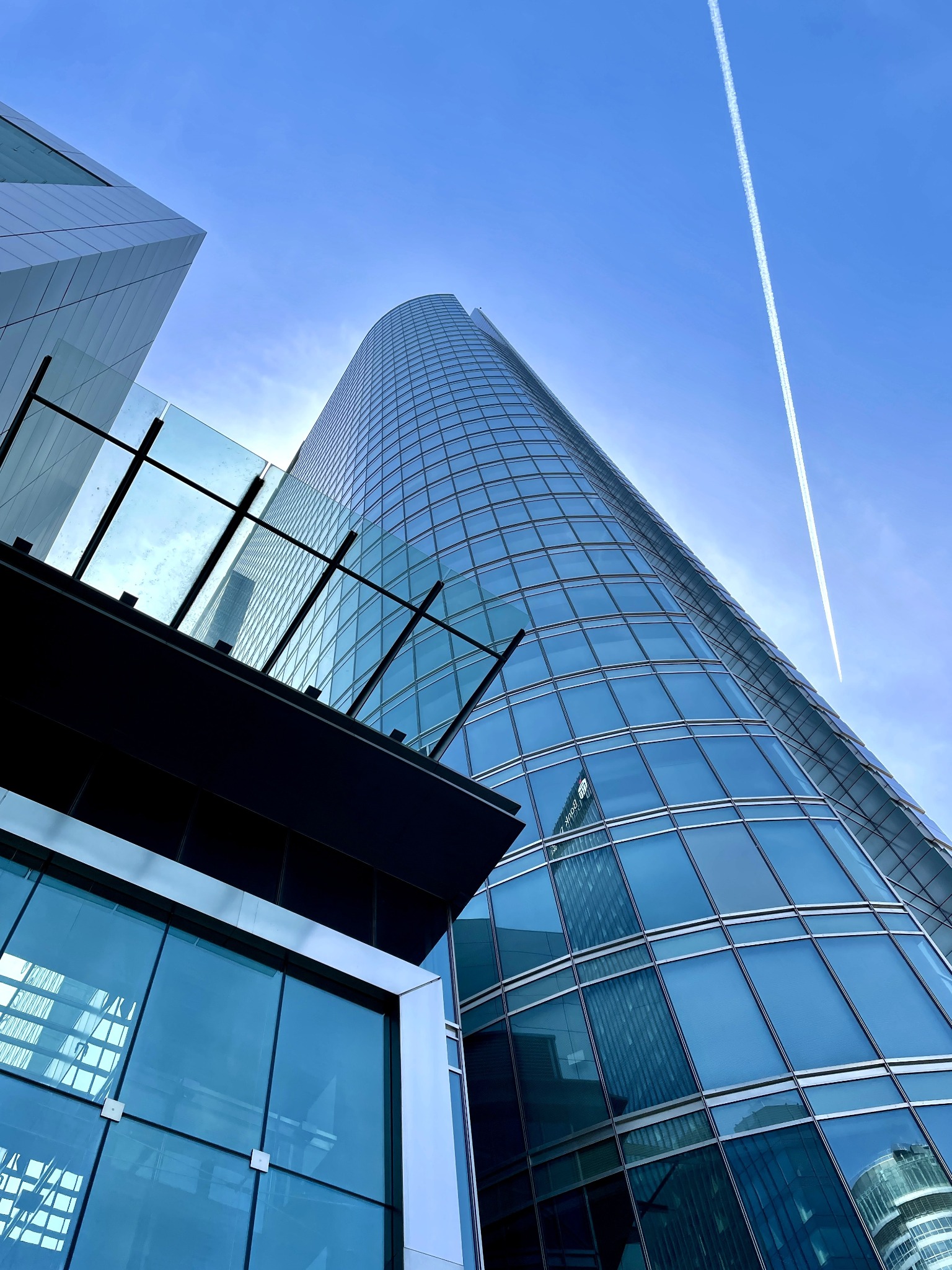 Wysoki, nowoczesny biurowiec z zaokrągloną fasadą odbijający inne budynki, widziany z dołu na tle błękitnego nieba z śladem kondensacyjnym samolotu.