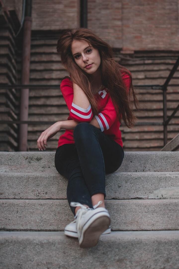 Portret młodej kobiety z rudymi włosami siedzącej na betonowych schodach w czerwonej bluzie z białymi paskami na tle ceglanej ściany.