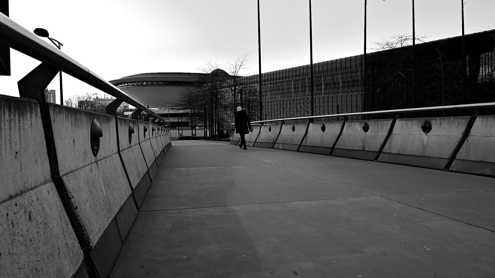 Czarno-białe zdjęcie kobiety idącej betonowym mostem z widokiem na nowoczesny budynek z ażurową fasadą i stadion w tle.