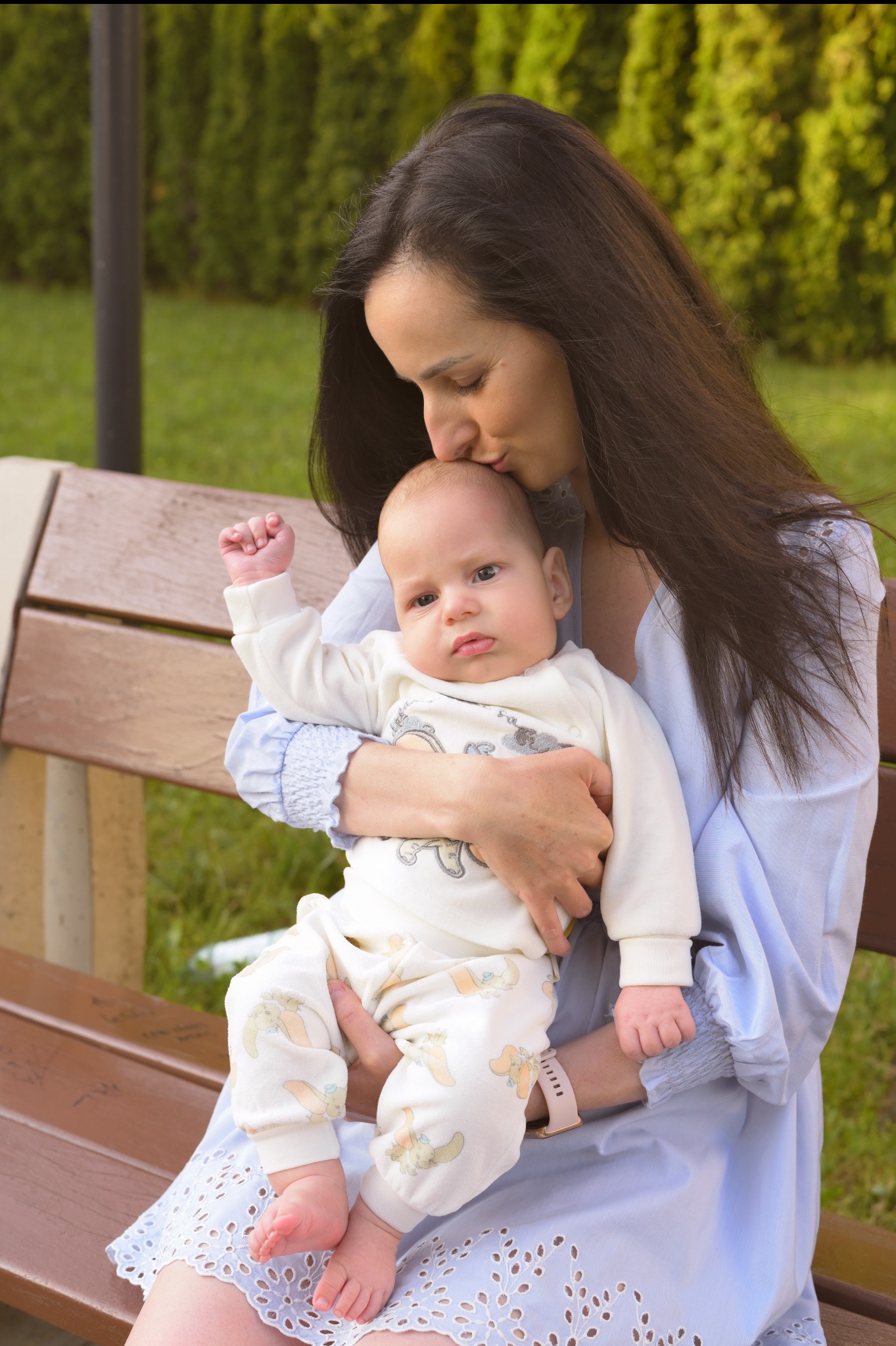 Matka całuje w głowę niemowlę ubrane w białe ubranko z króliczkami, siedząc na ławce w parku, w tle zieleń. Delikatne, naturalne światło.